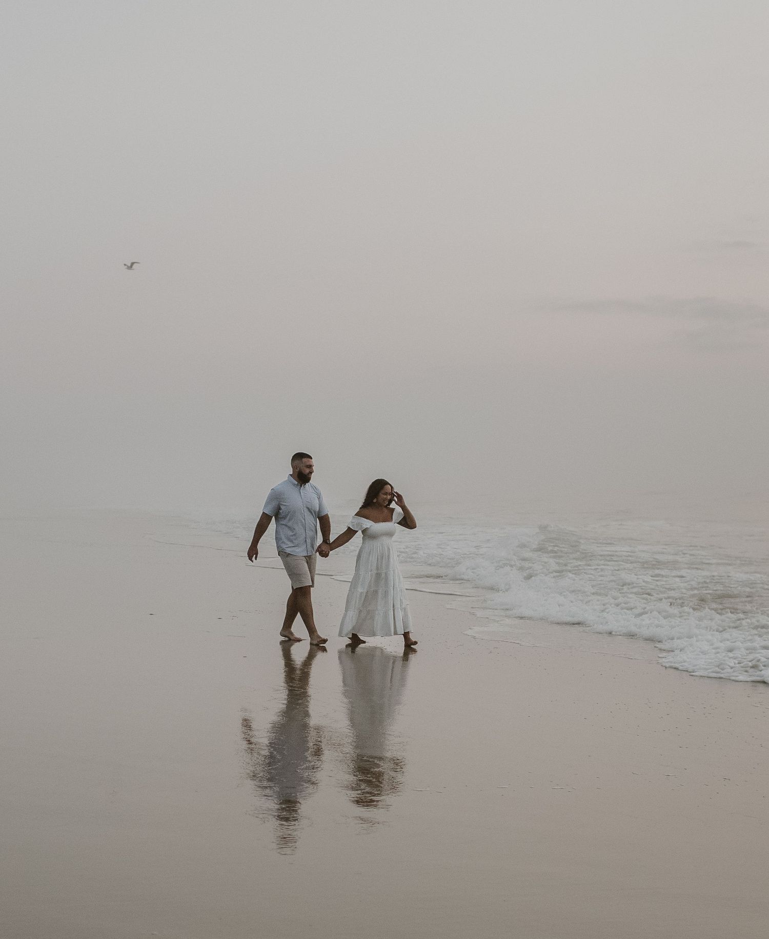 Couple holding hands walking along wet shoreline on a misty overcast beach with reflections.