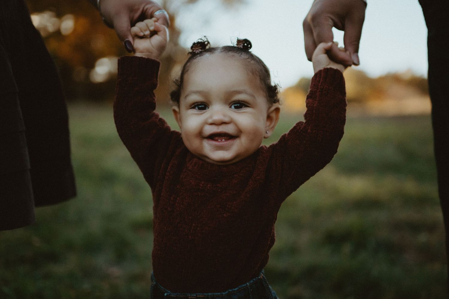 Happy toddler girl in red sweater standing with arms raised held by parents outdoors.