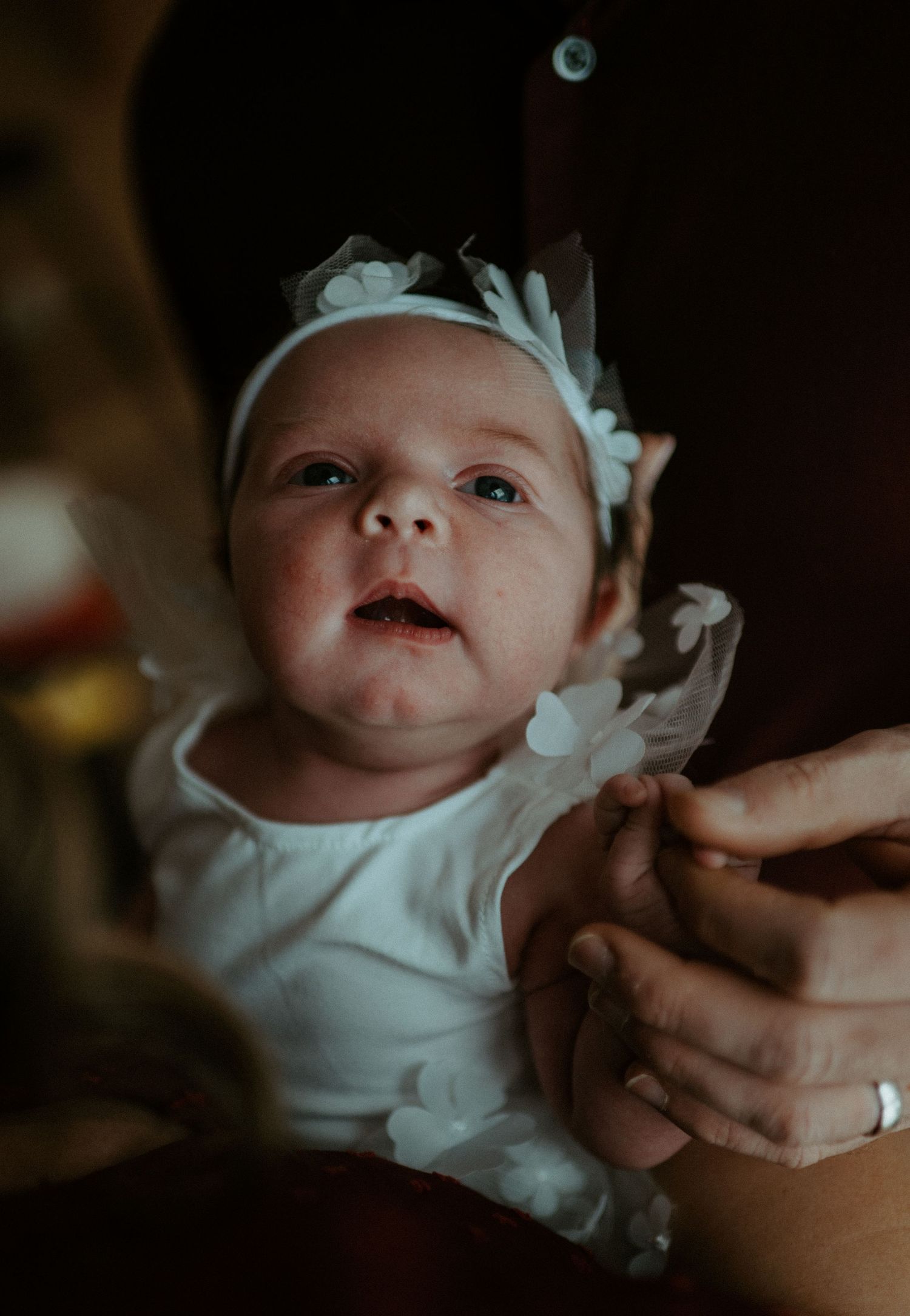 Adorable newborn baby girl in white floral headband and lace outfit looking up at the camera.