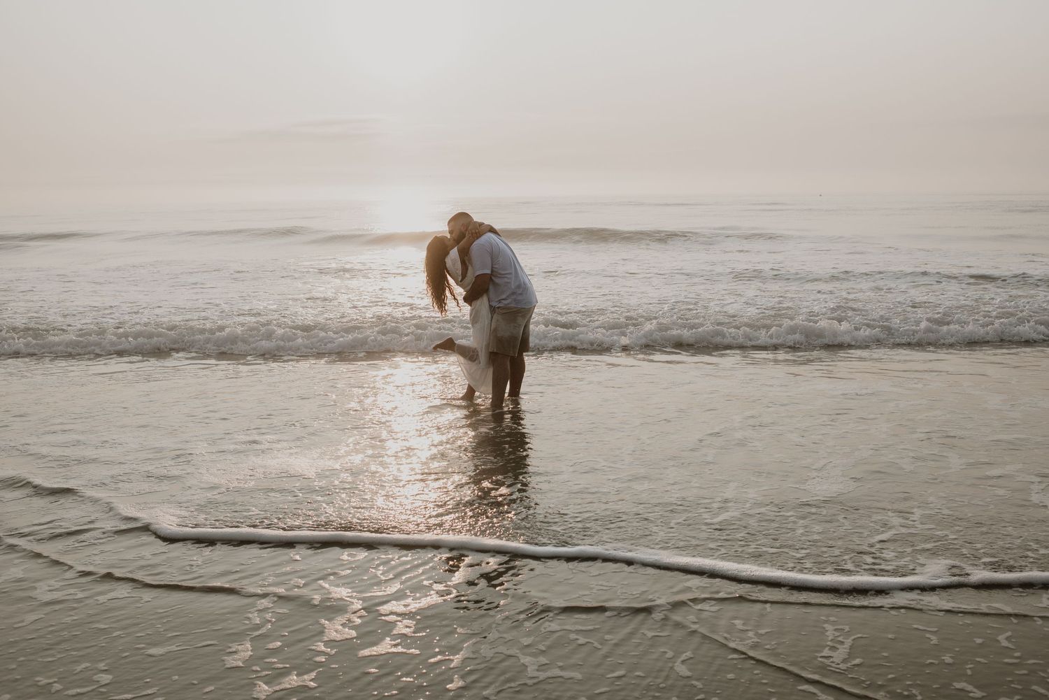 Couple sharing a tender moment standing in shallow ocean water at sunset on the beach.