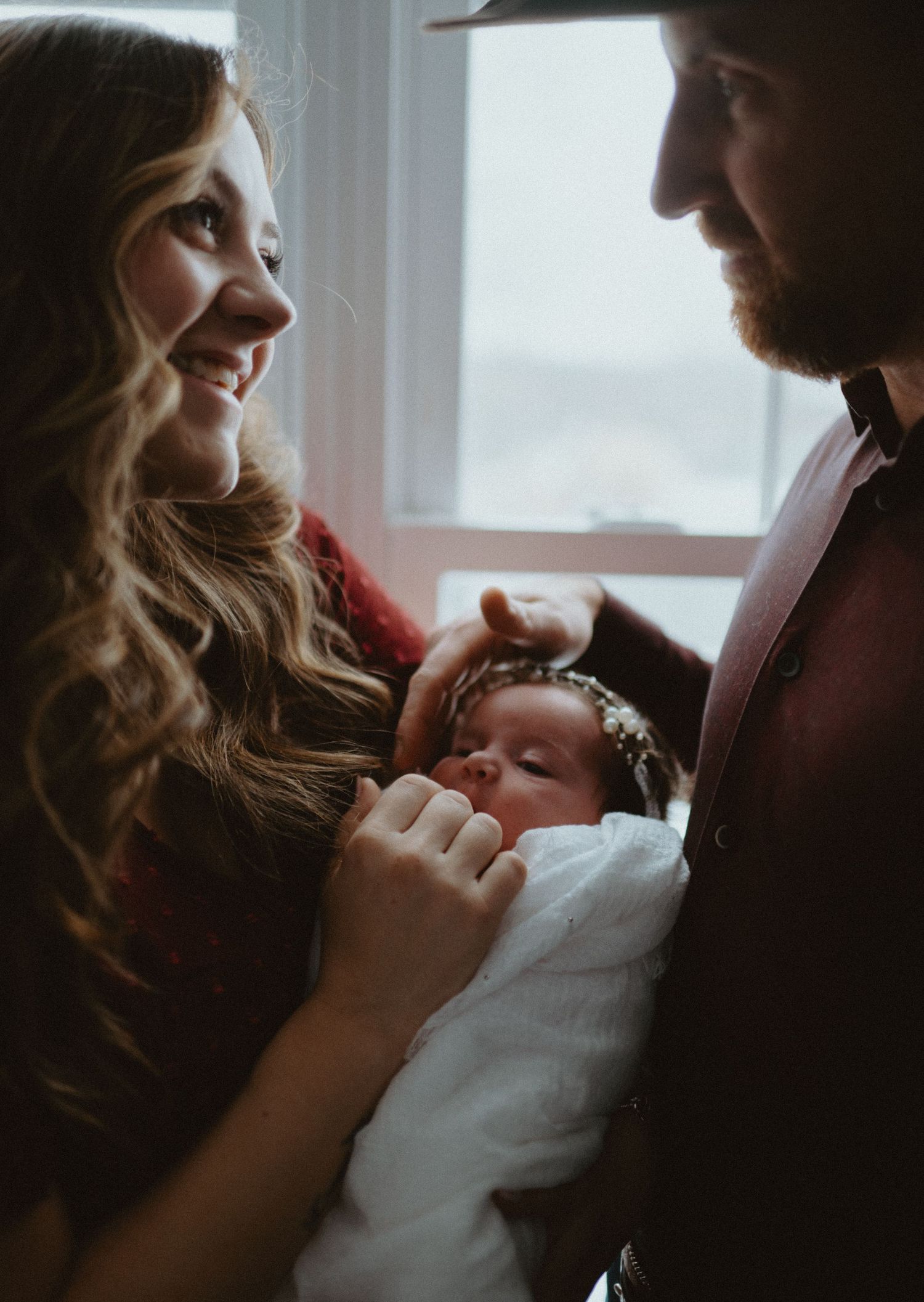 Happy parents smiling at their sleeping newborn baby wrapped in white by a bright window.