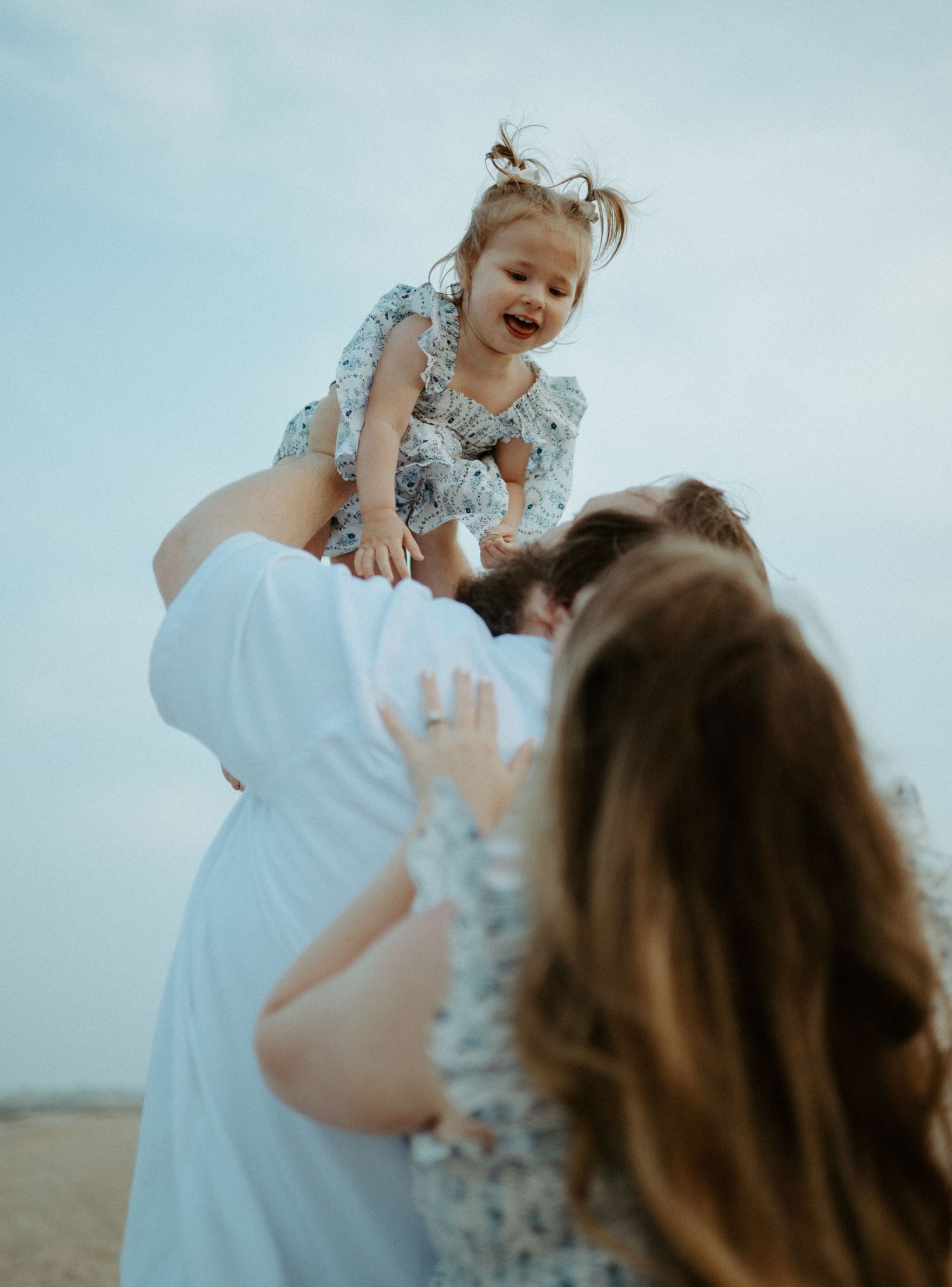 Happy toddler girl laughing as parents lift her up playfully on a sandy beach at dusk.