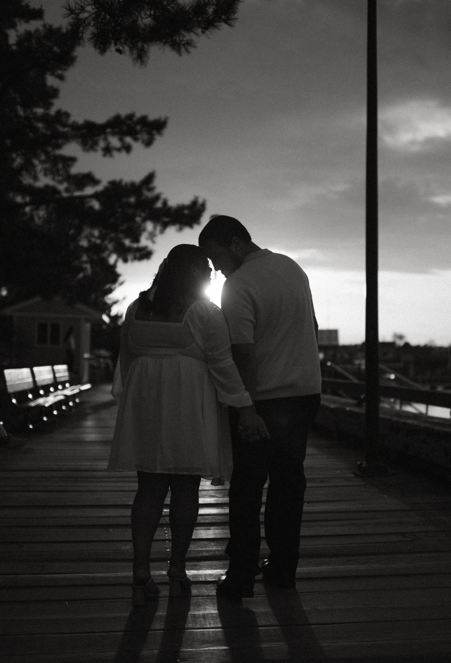 Black and white silhouette of a couple touching foreheads on a boardwalk with sun glowing behind them.