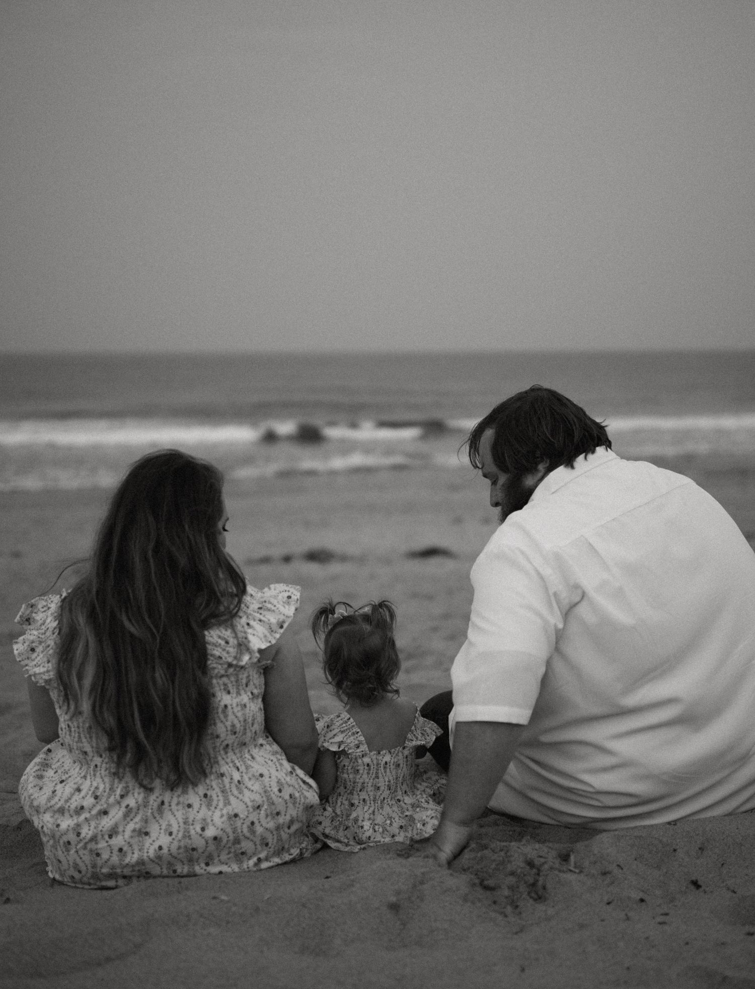 Black and white photo of a family of three sitting together on the beach facing the ocean.