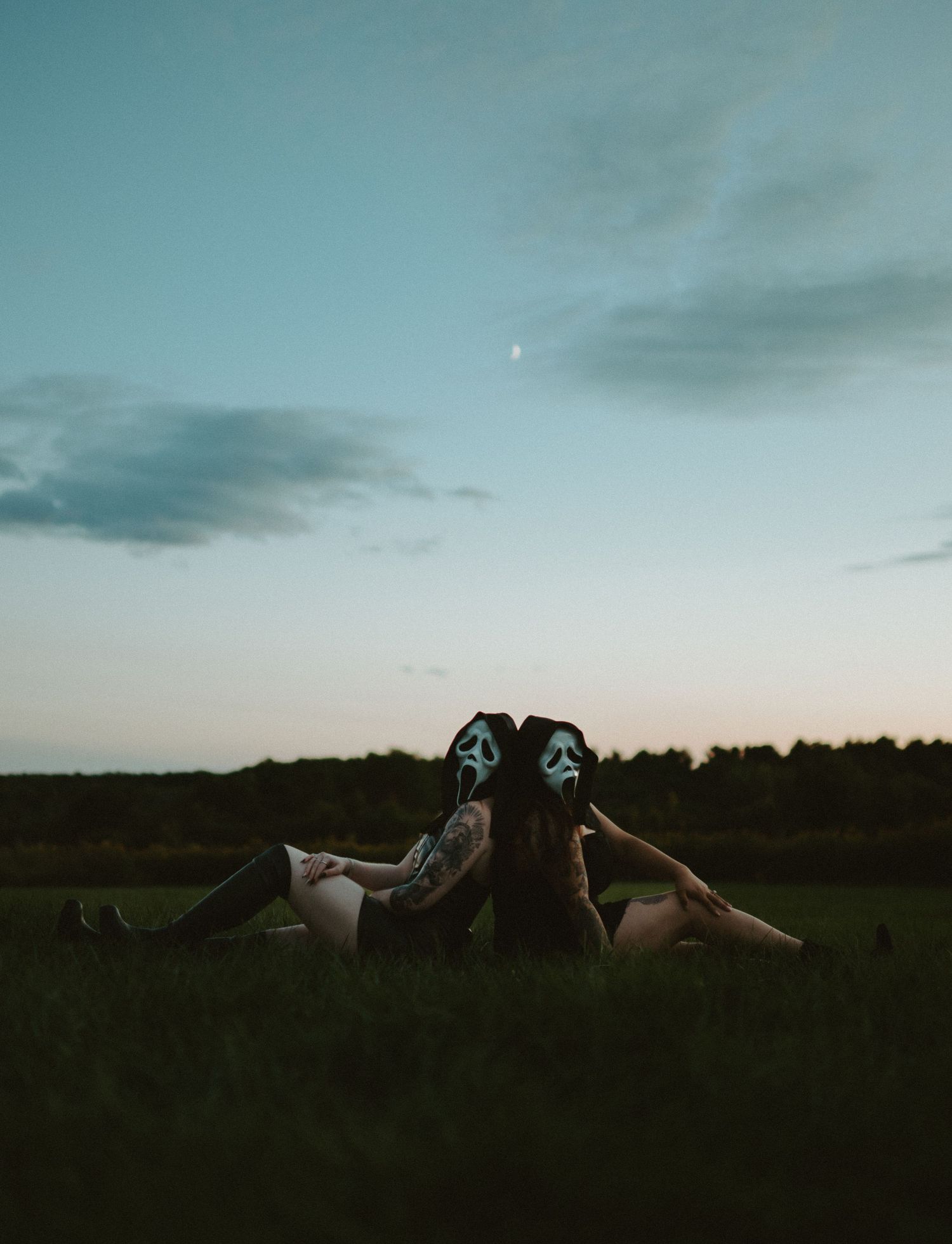 Two people in Ghostface Halloween masks sitting back-to-back on grass under a twilight sky.