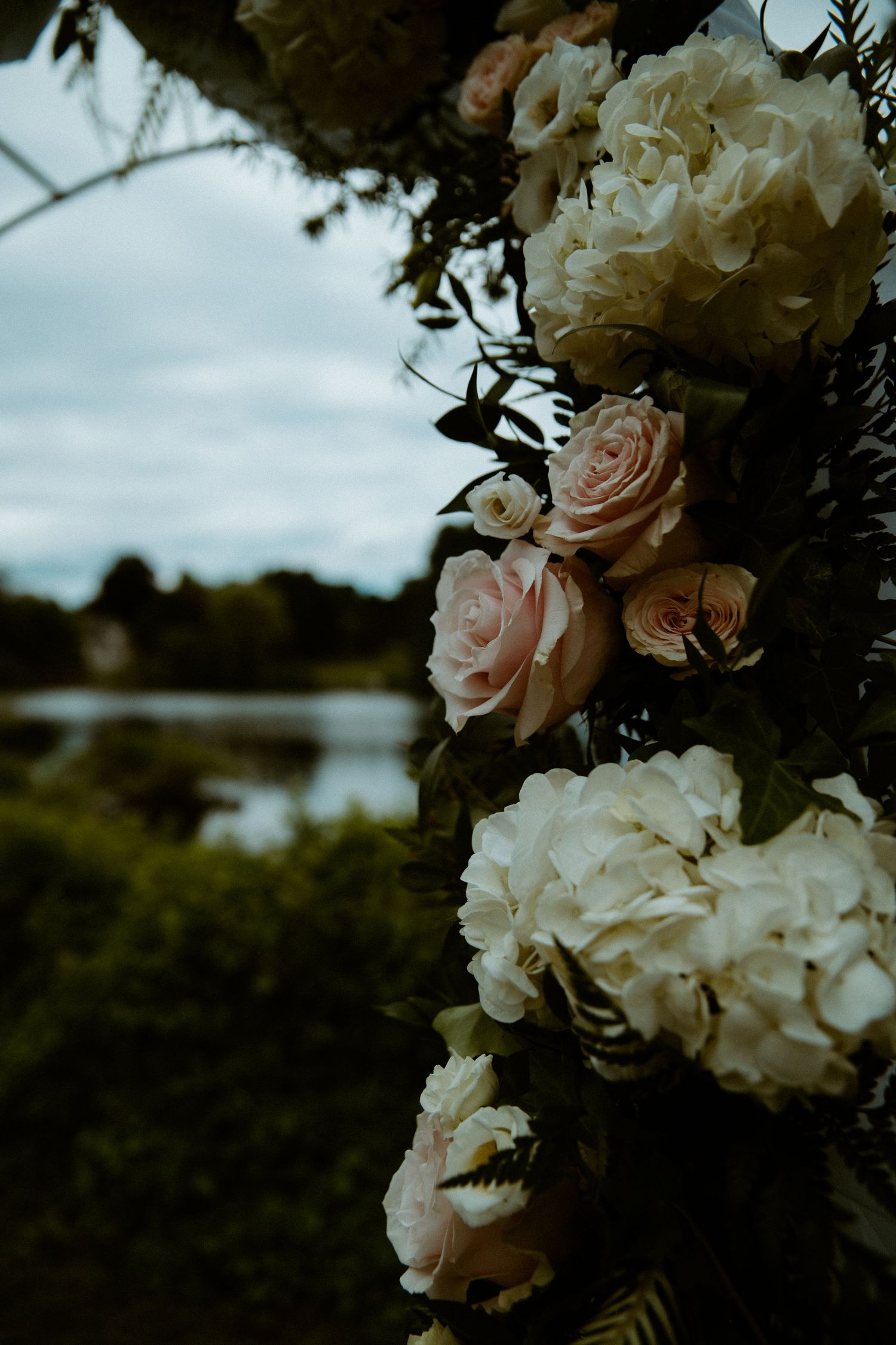 Elegant wedding arch adorned with blush pink roses and white hydrangeas against a serene waterfront backdrop.