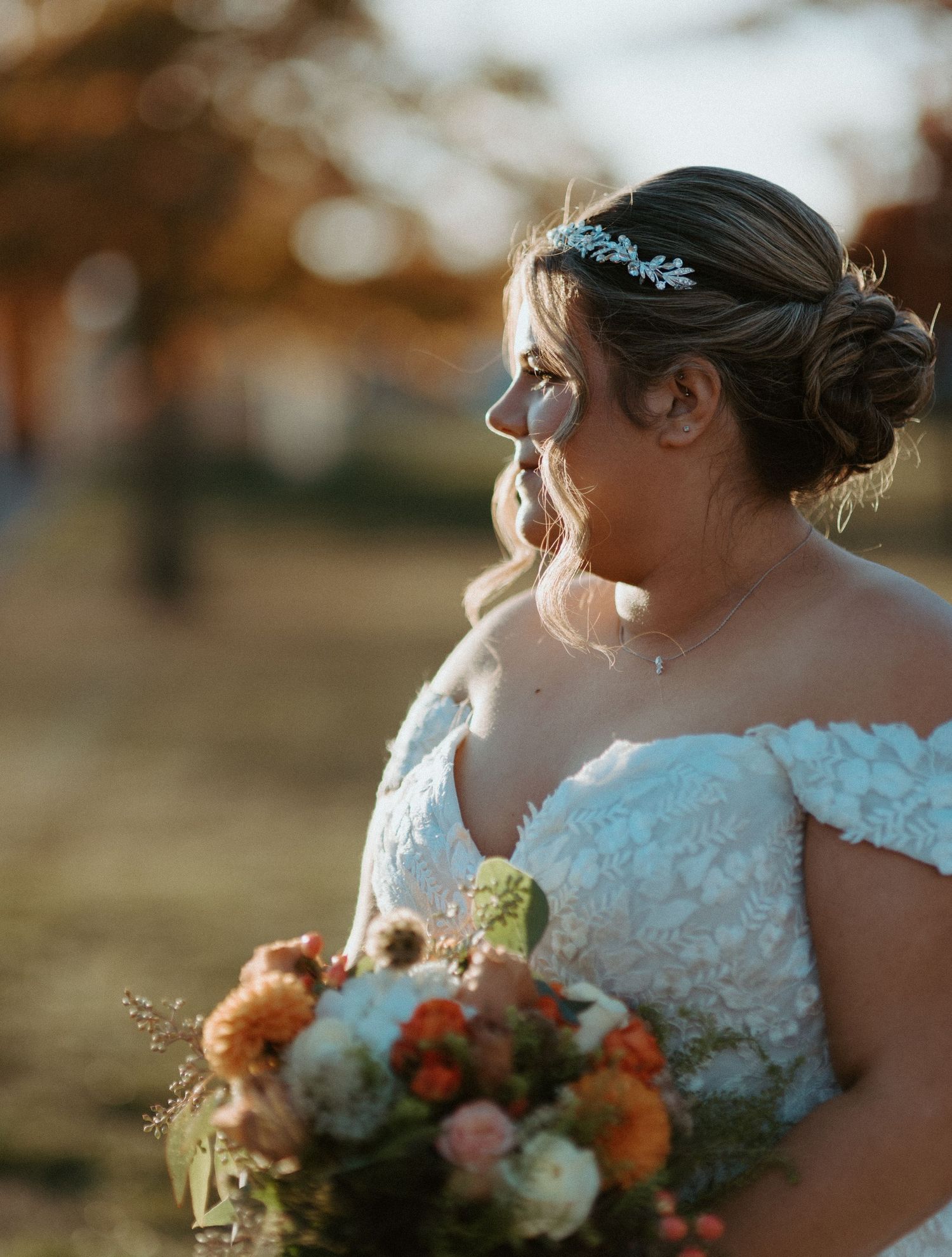 Bride in off-shoulder lace gown holding colorful bouquet with floral headpiece in golden sunlight.