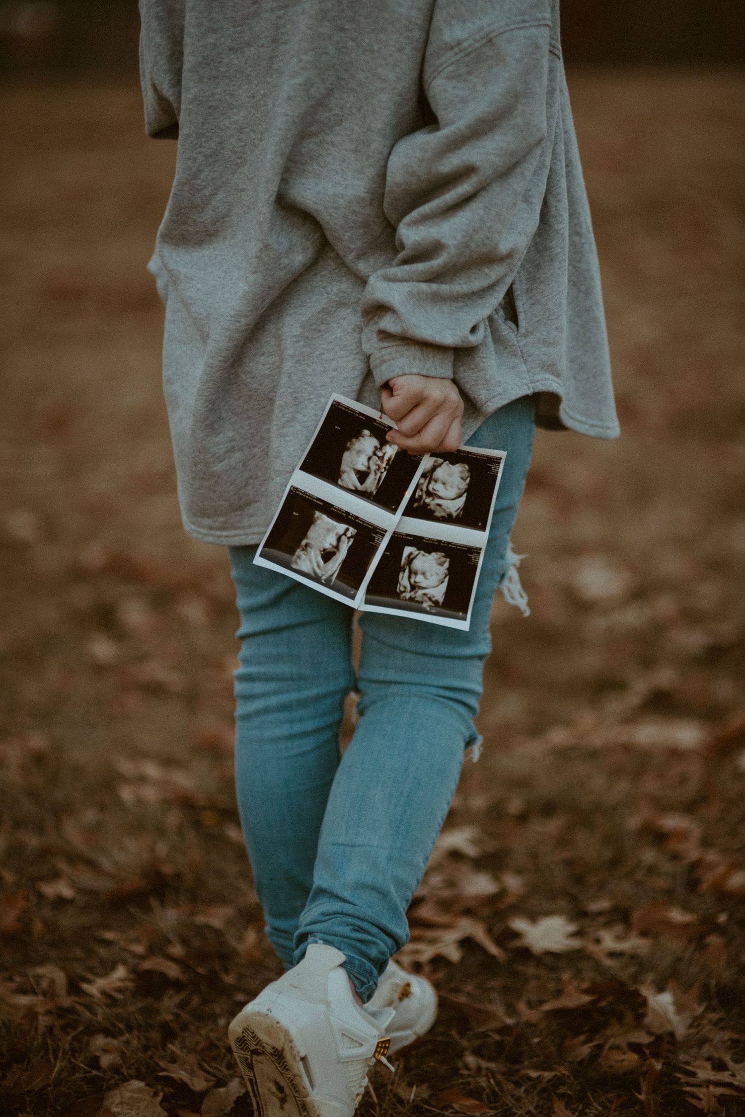 Close-up of pregnant woman holding ultrasound photos while walking on fallen autumn leaves.