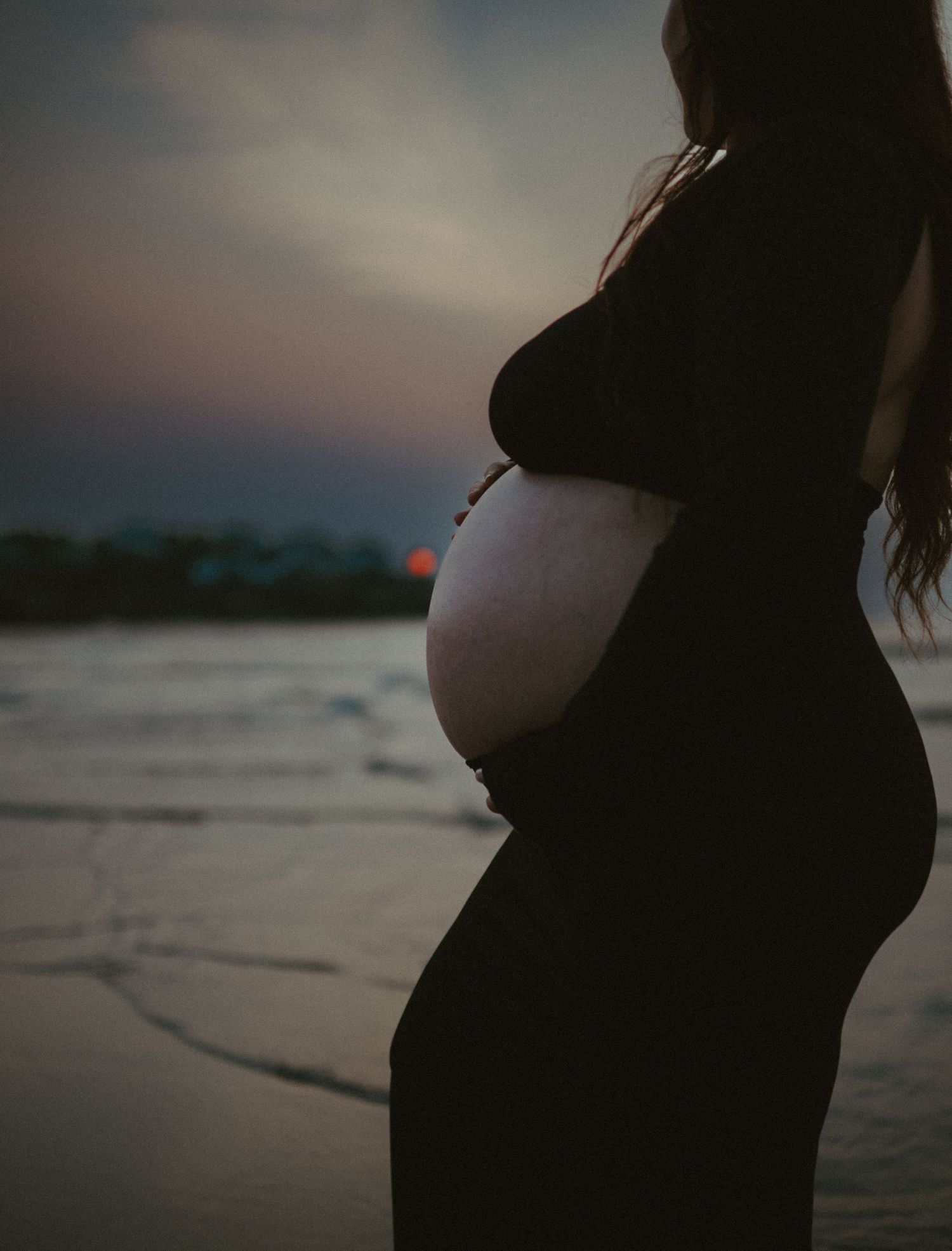Close-up silhouette of pregnant woman in black dress cradling bare baby bump at sunrise on beach.