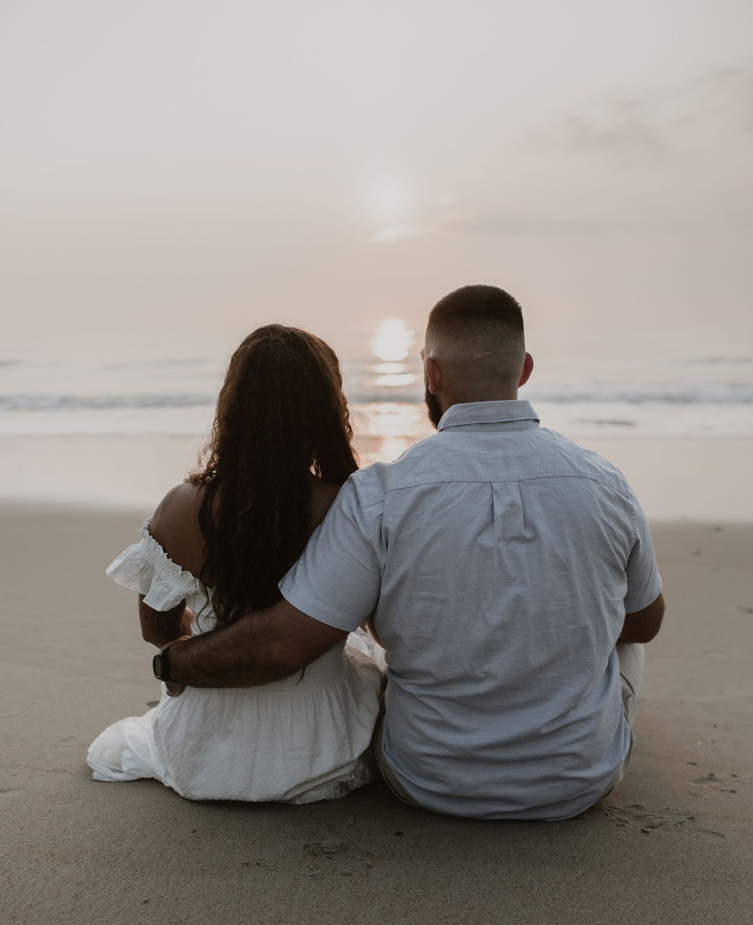 Couple sitting together on beach watching sunset over ocean waves, viewed from behind.