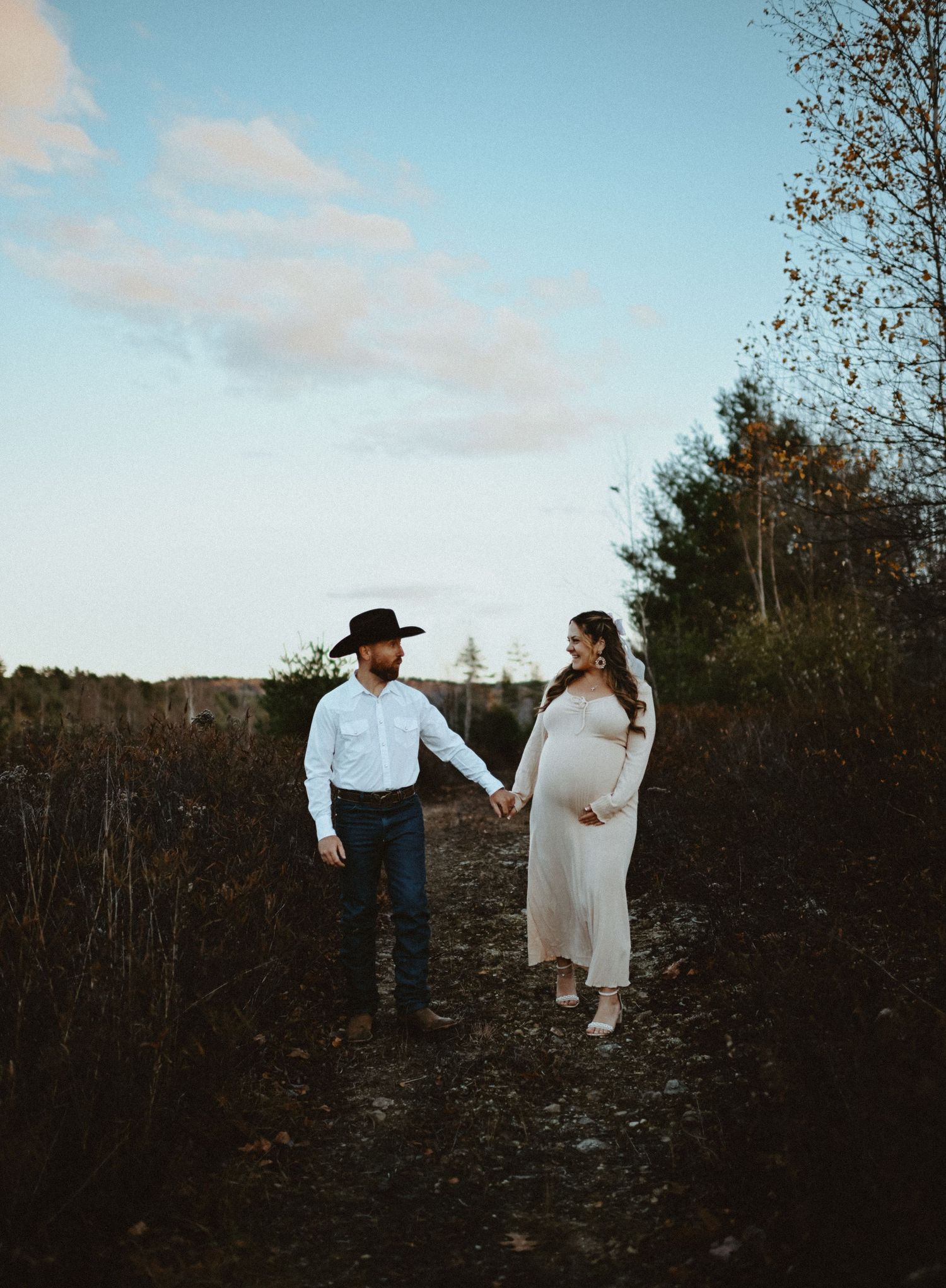 Pregnant couple holds hands on rural path, man in cowboy hat and woman in cream maternity gown.
