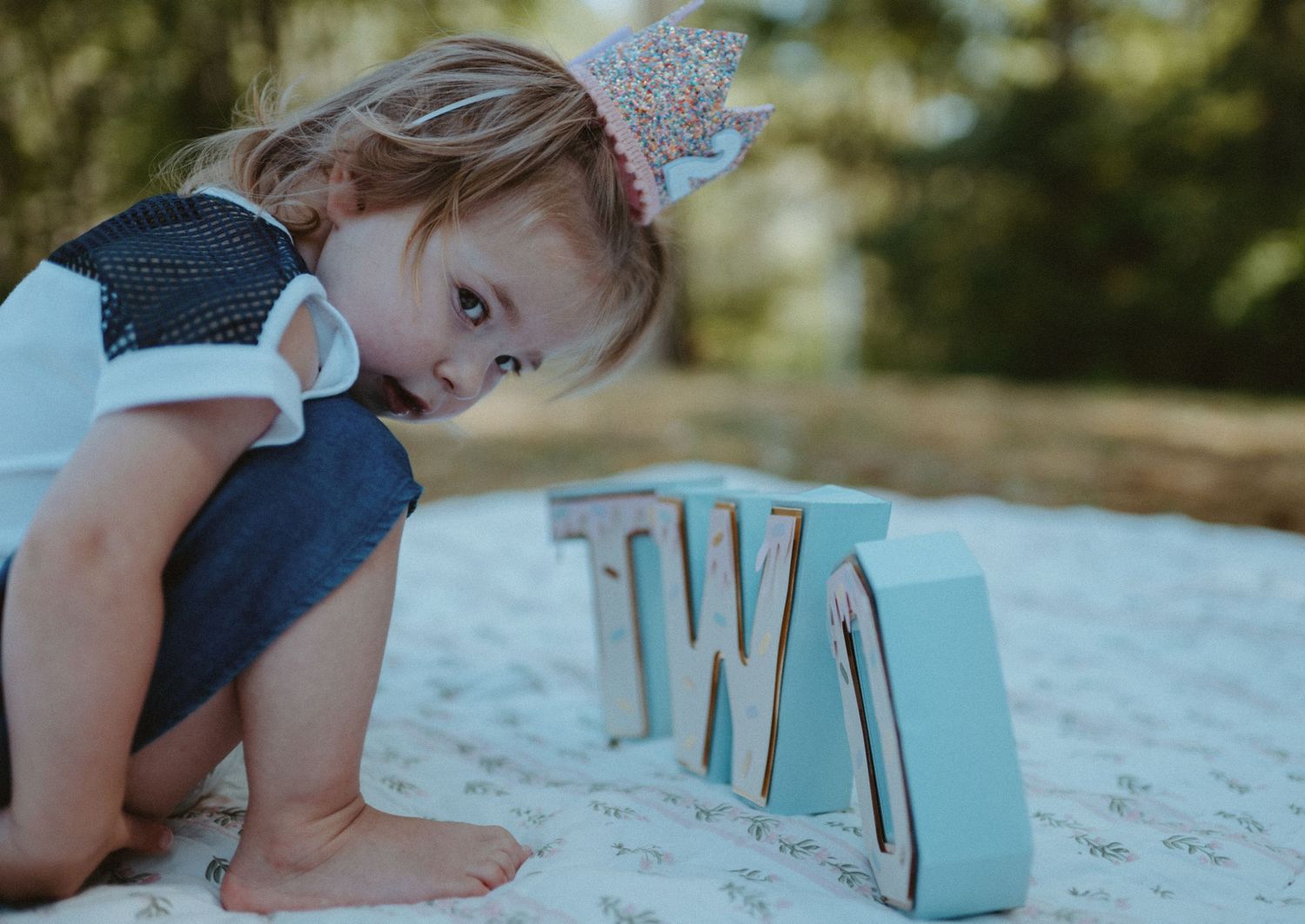 Curious toddler wearing a birthday crown crawling toward TWO letter signs during outdoor photo session.