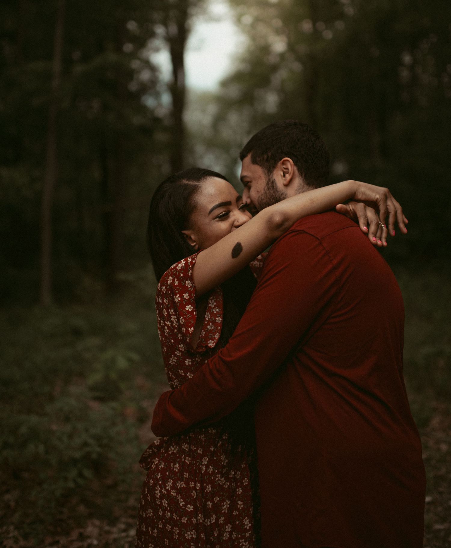 Couple sharing a tender embrace in a moody forest setting, both dressed in deep red tones.