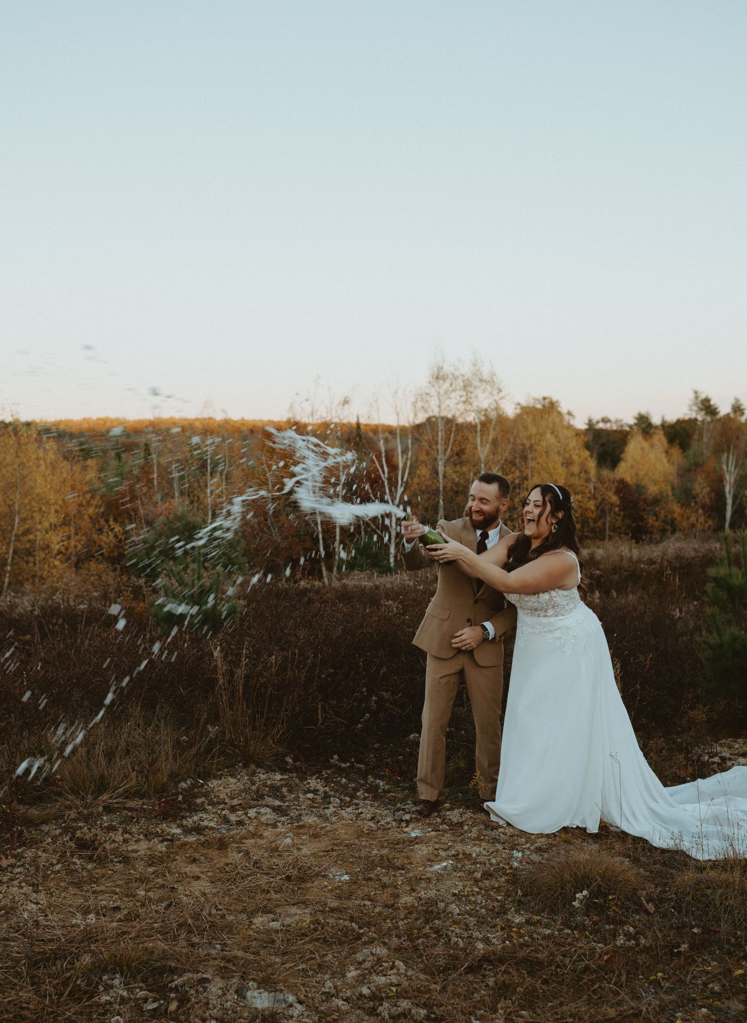 Smiling couple popping champagne bottle outdoors surrounded by colorful autumn foliage.