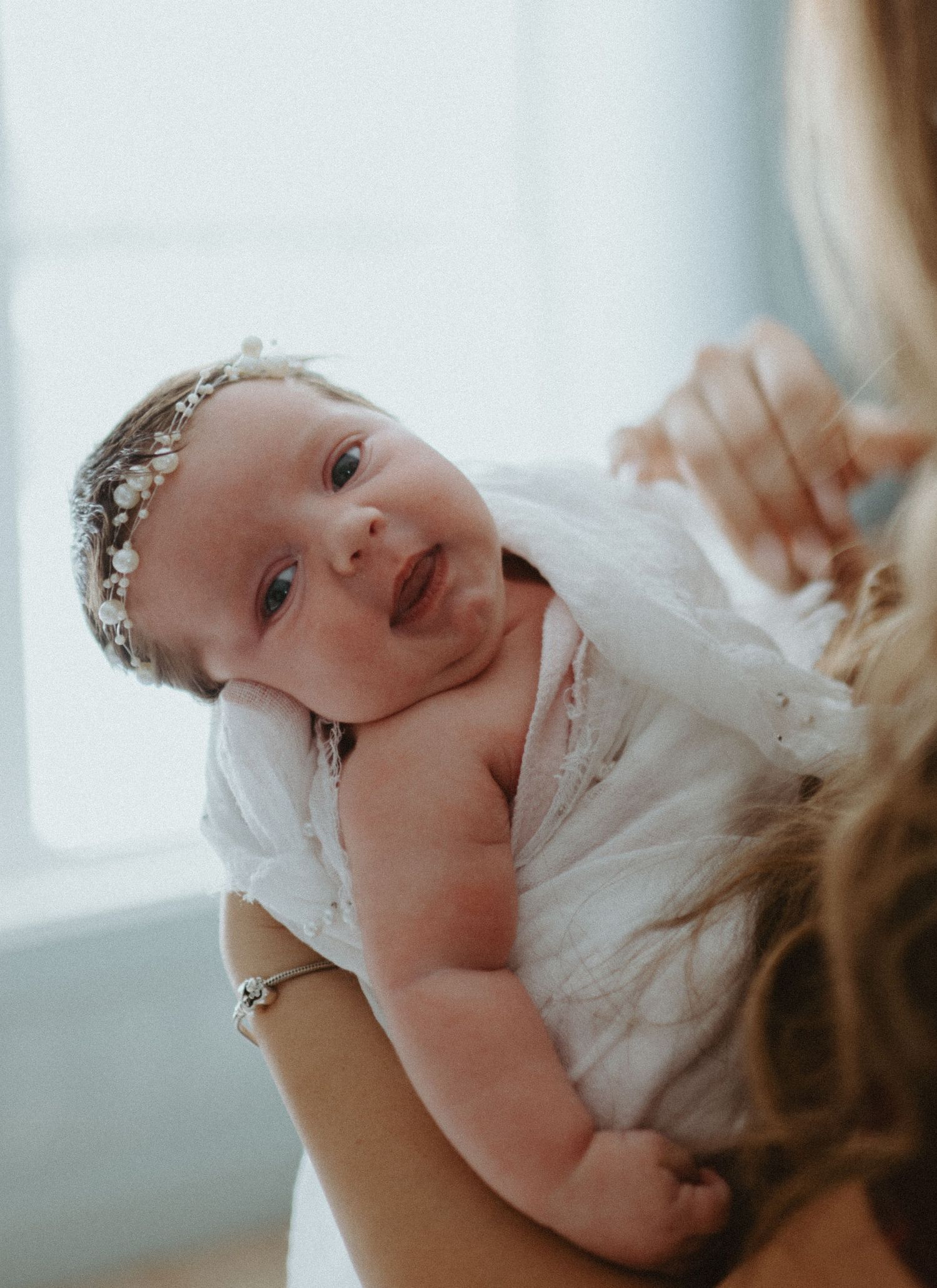 Alert newborn baby with pearl headband being held up by mother in soft natural light.