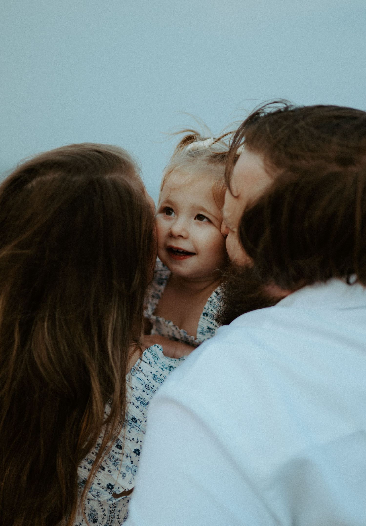 Two parents lovingly kiss their smiling toddler girl on the cheeks in a tender family portrait.