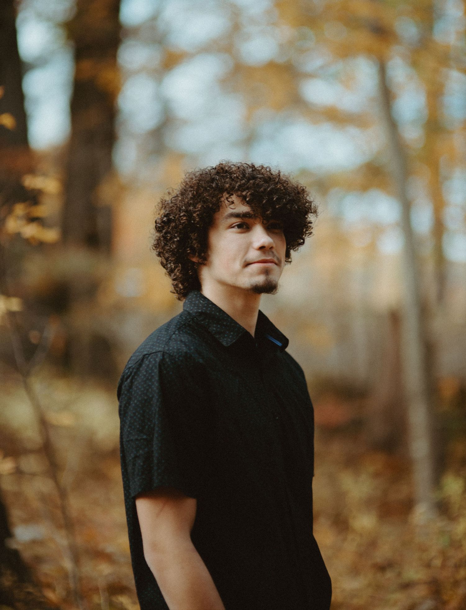 Young man with curly hair in black shirt standing in autumn forest, looking over shoulder.
