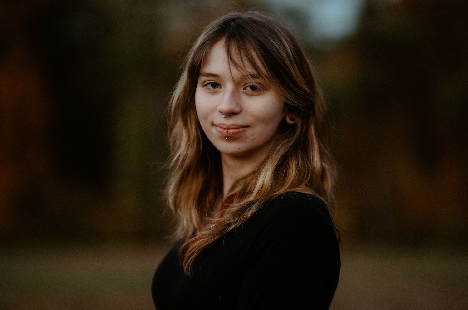 Young woman with wavy hair smiling softly outdoors in a black top against autumn foliage bokeh background.