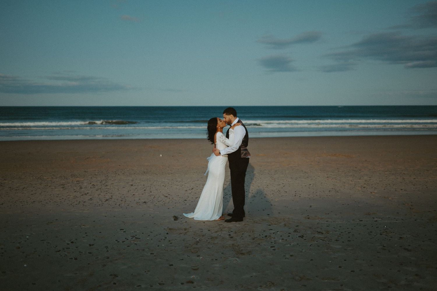 Bride and groom share a kiss on a serene beach at dusk with ocean waves in the background.