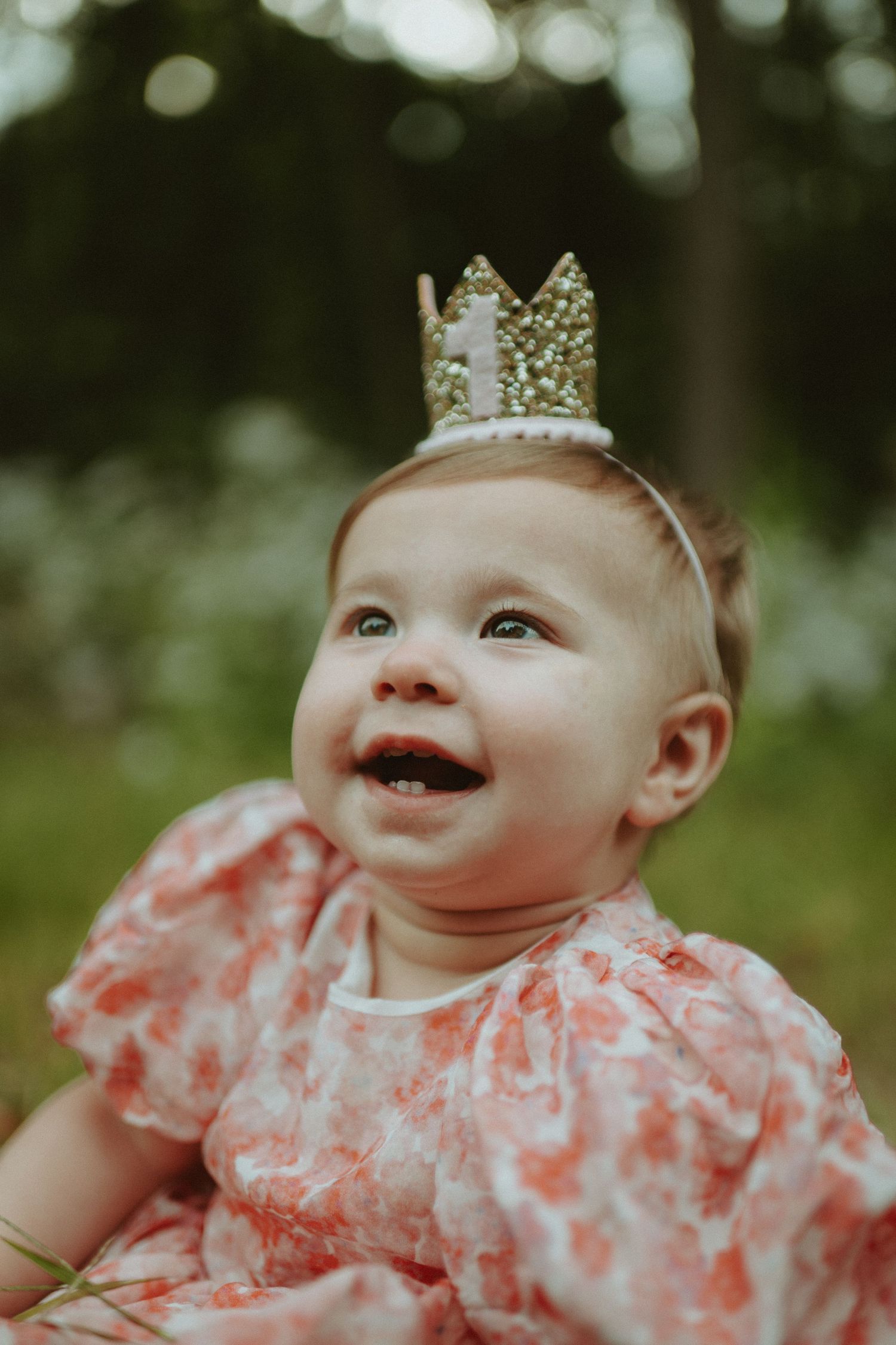 Smiling baby girl wearing gold crown headband in pink floral dress outdoors.