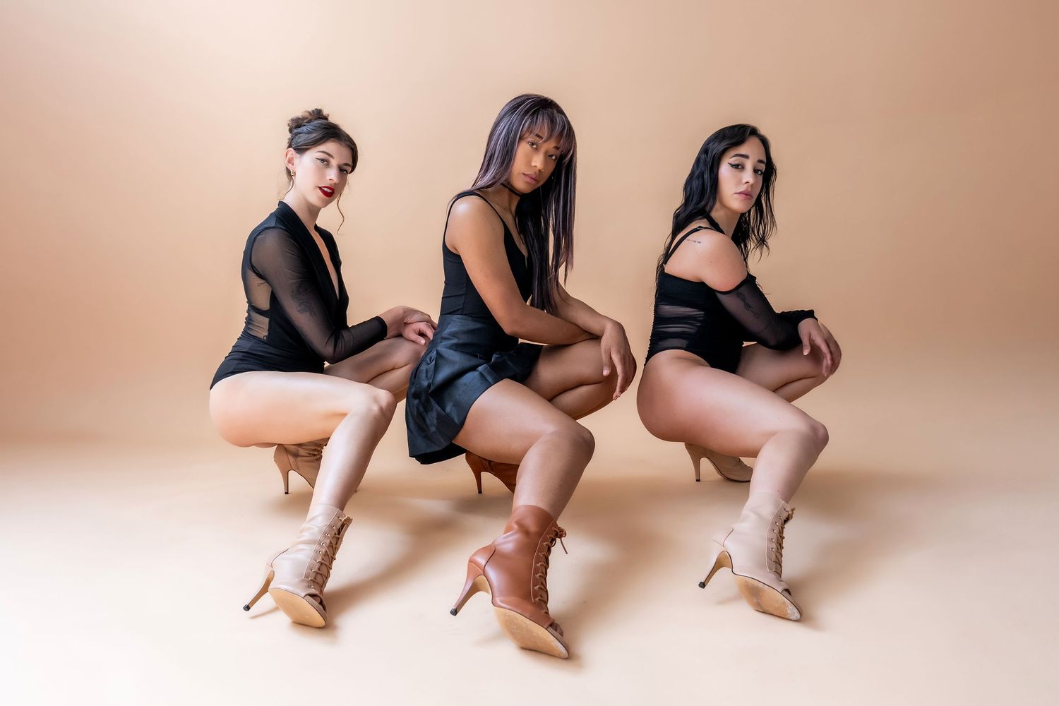 Three women in black outfits and heels pose confidently in a studio with a beige background.