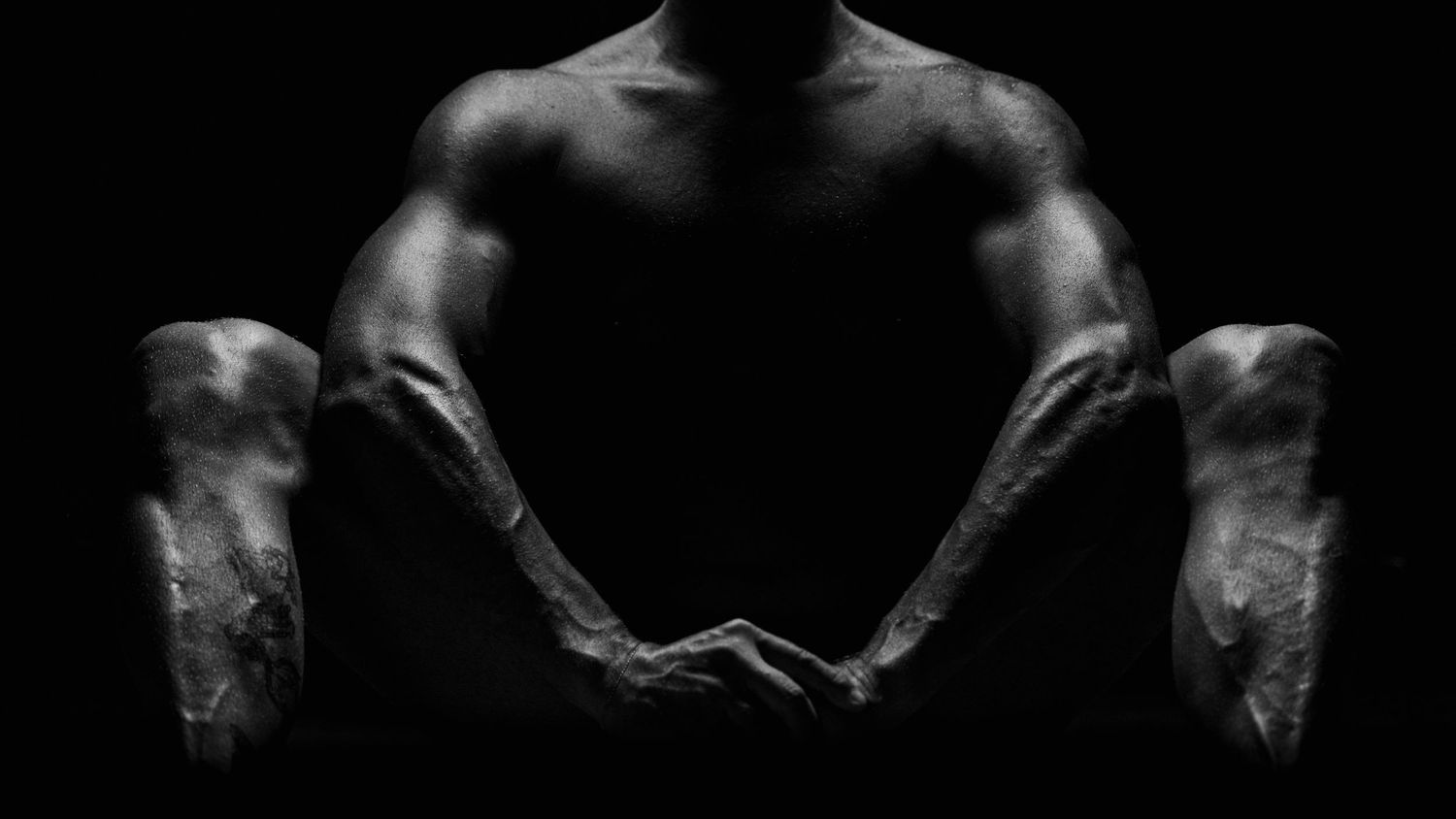 Black and white dramatic close-up of a muscular male torso and veined forearms on dark background.