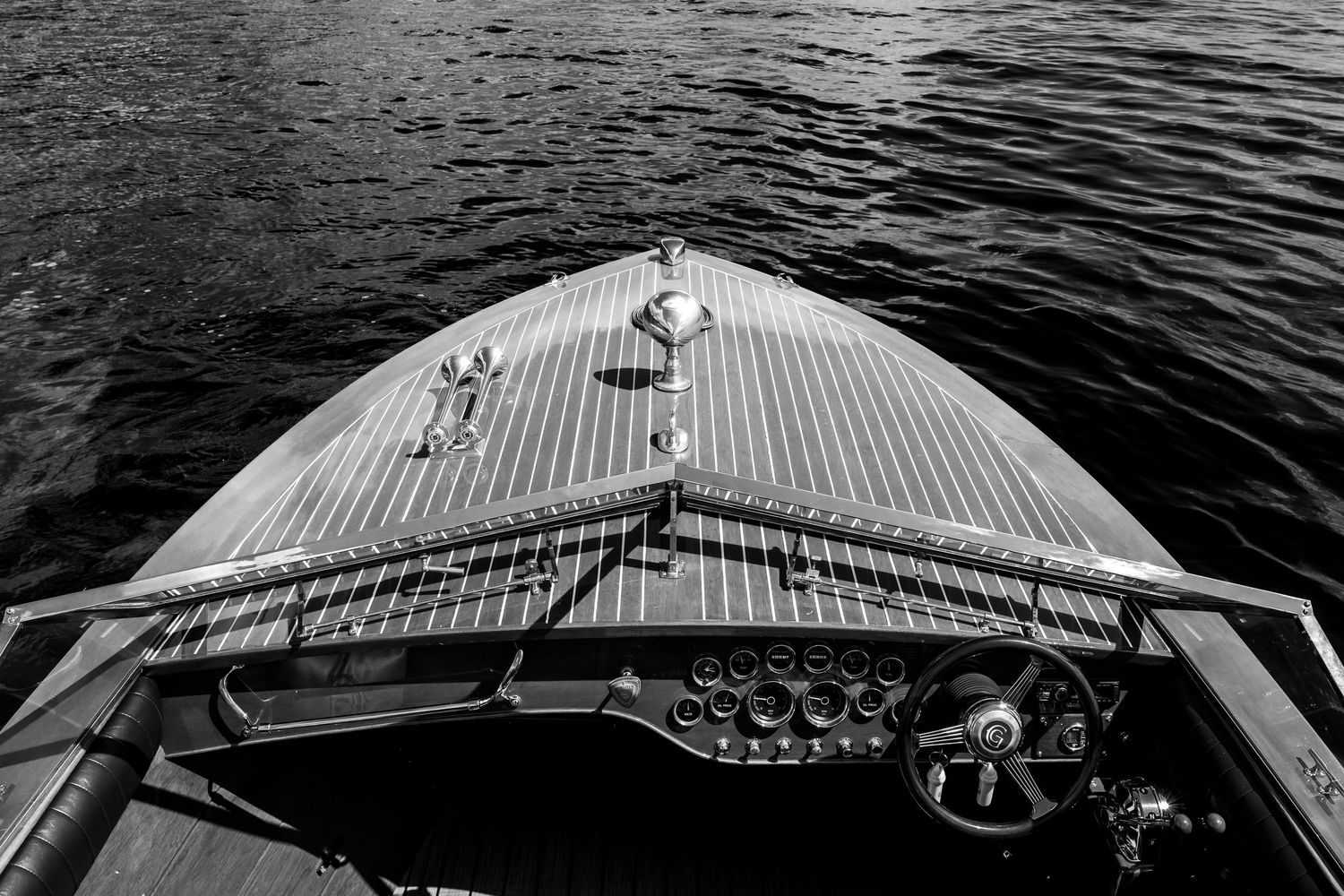 Black and white photo of a classic wooden speedboat bow with steering wheel on calm water.