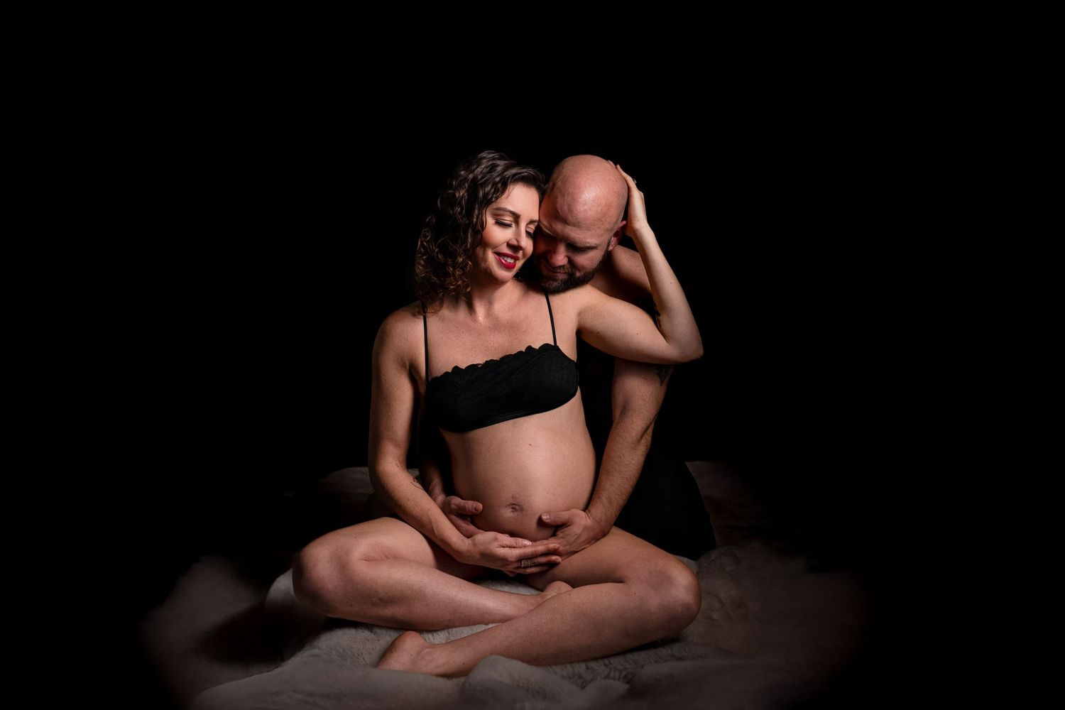 Couple expecting a baby, man embracing pregnant woman's belly, sitting together in a dark studio setting.