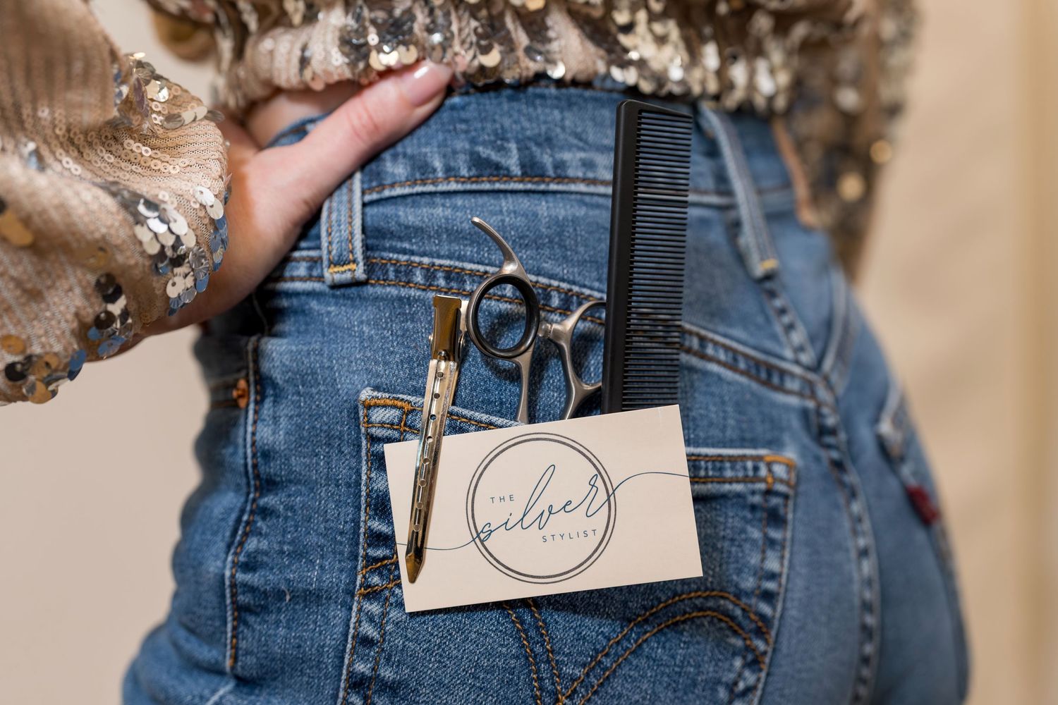 Hairstylist holding scissors and a business card in denim jeans back pocket, wearing a sequin top.