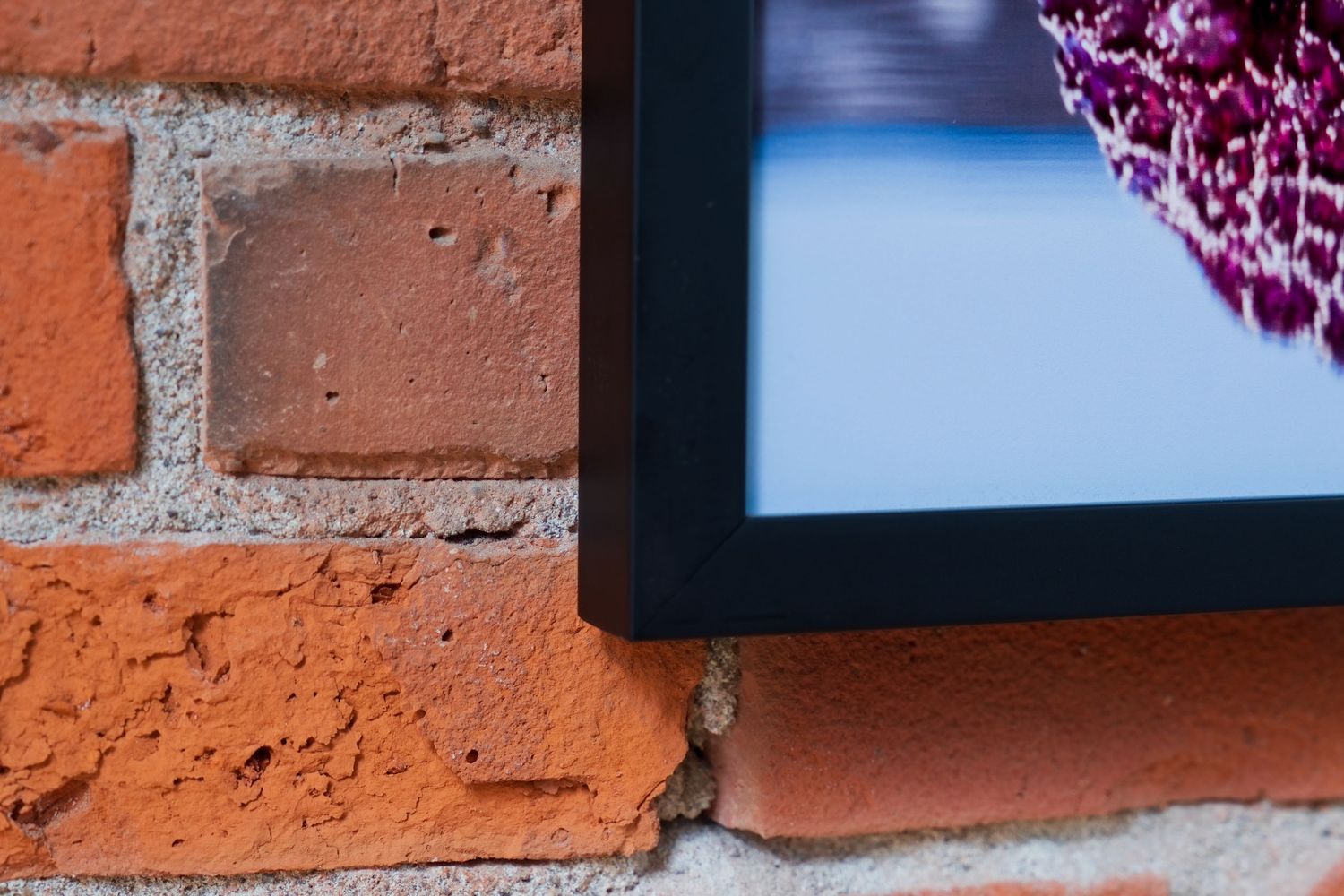 A black picture frame with floral art hangs on a rustic red brick wall, showcasing texture and contrast.