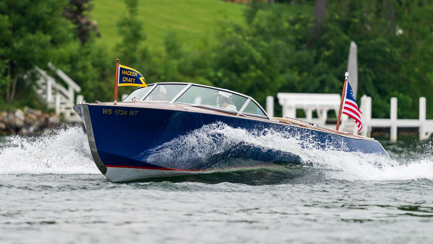A classic blue wooden speedboat with an American flag cuts through lake waters, creating white spray waves.