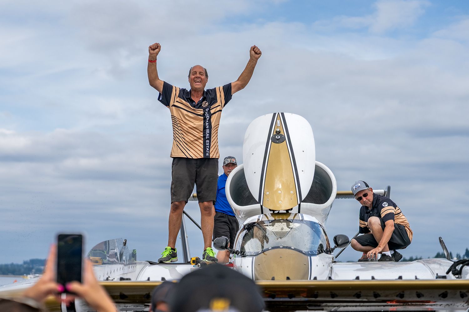 A triumphant man raises both arms in victory atop a racing hydroplane boat at a competitive watercraft event.