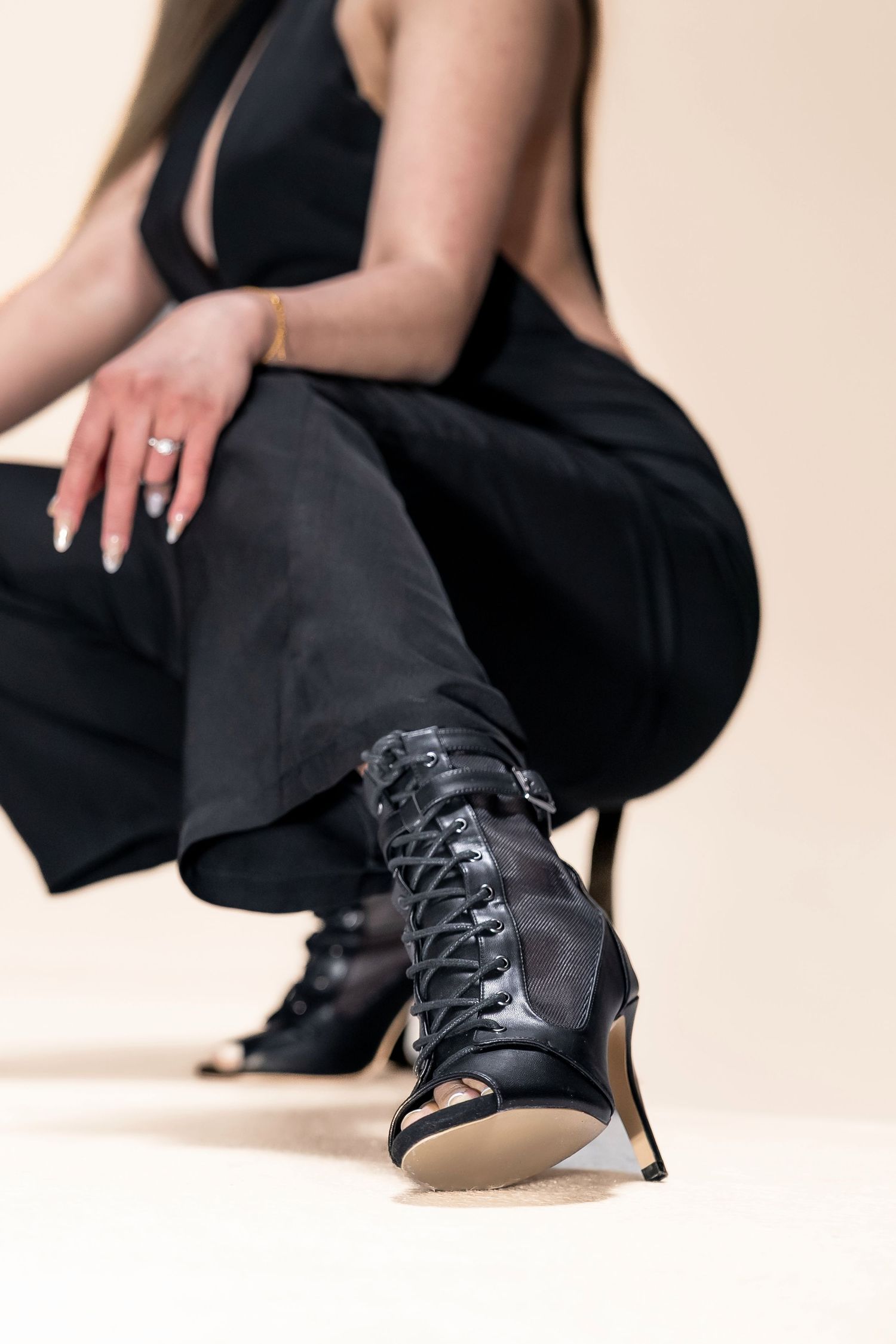 Woman in black outfit wearing lace-up black heeled boots, crouching on light background.