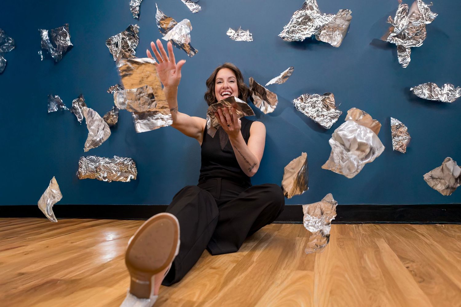 Woman sitting on floor tossing crumpled aluminum foil pieces against a blue wall in an art installation.