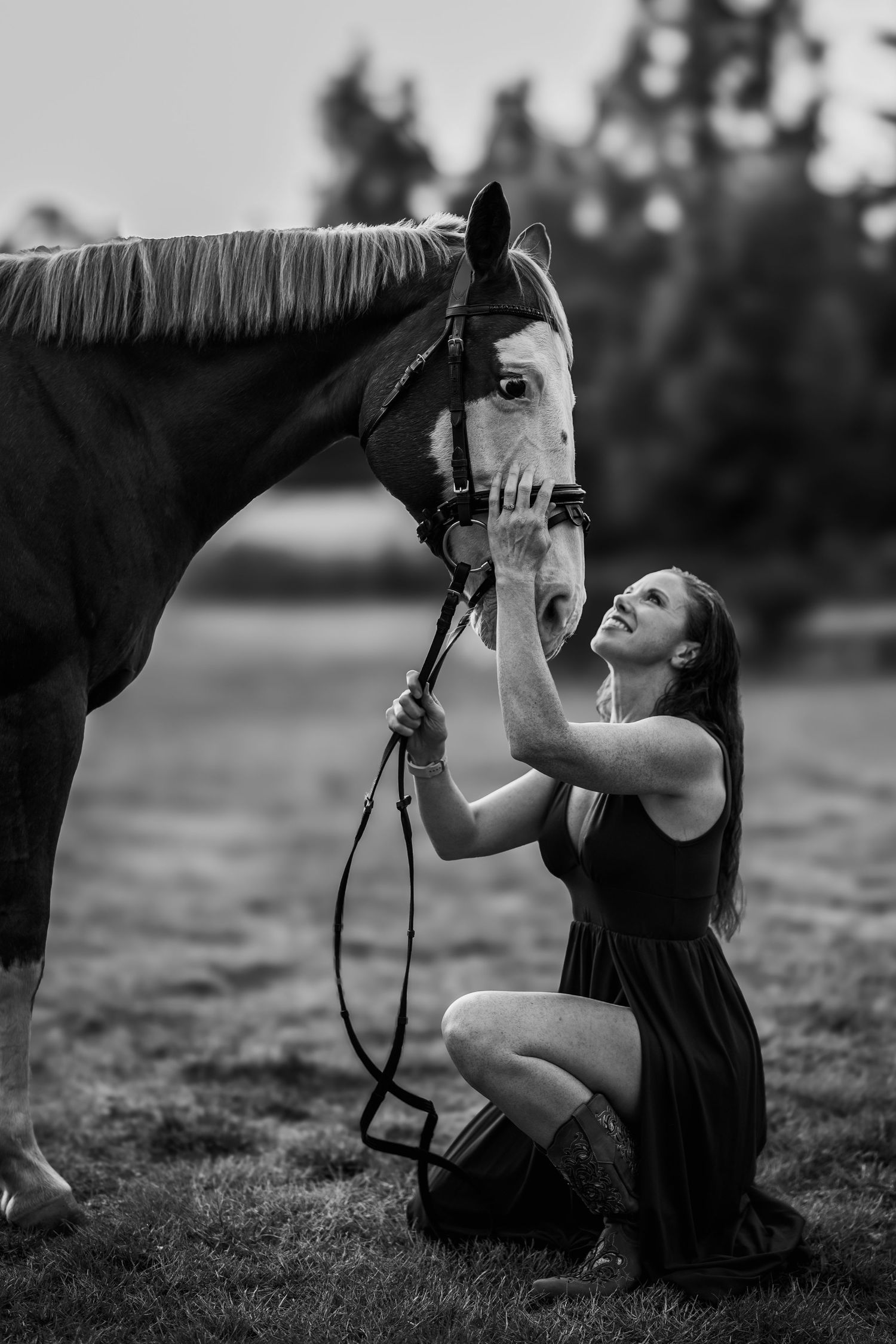 Black and white photo of a smiling woman kneeling beside a horse in an open field.