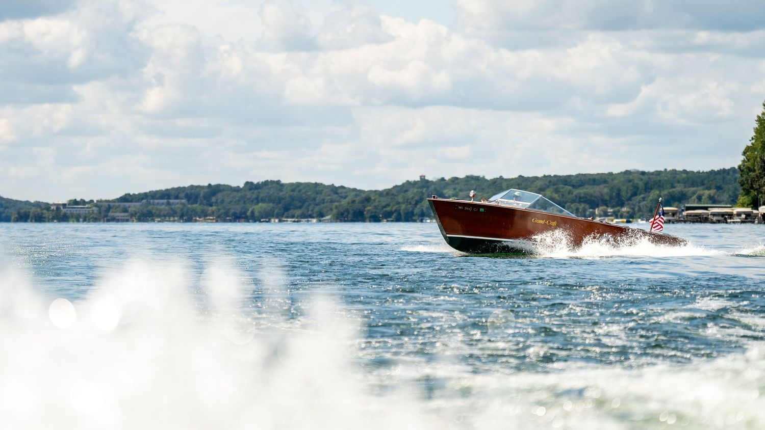 Classic wooden vintage speedboat speeding across a lake with white wake and cloudy sky.
