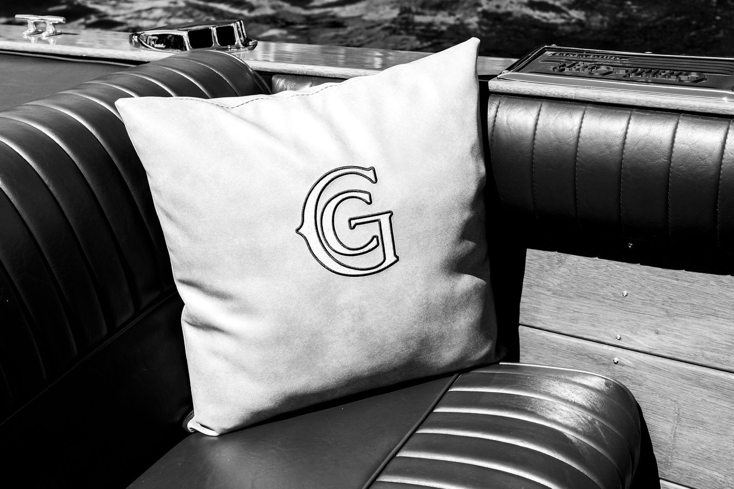 Black and white photo of a white decorative pillow with bold G monogram on classic black leather boat seat.