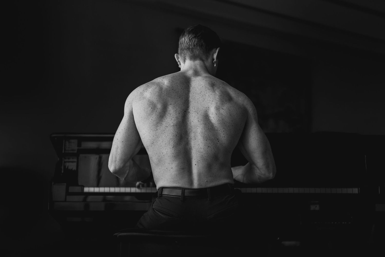 Black and white rear view of muscular shirtless man playing piano in dark room.