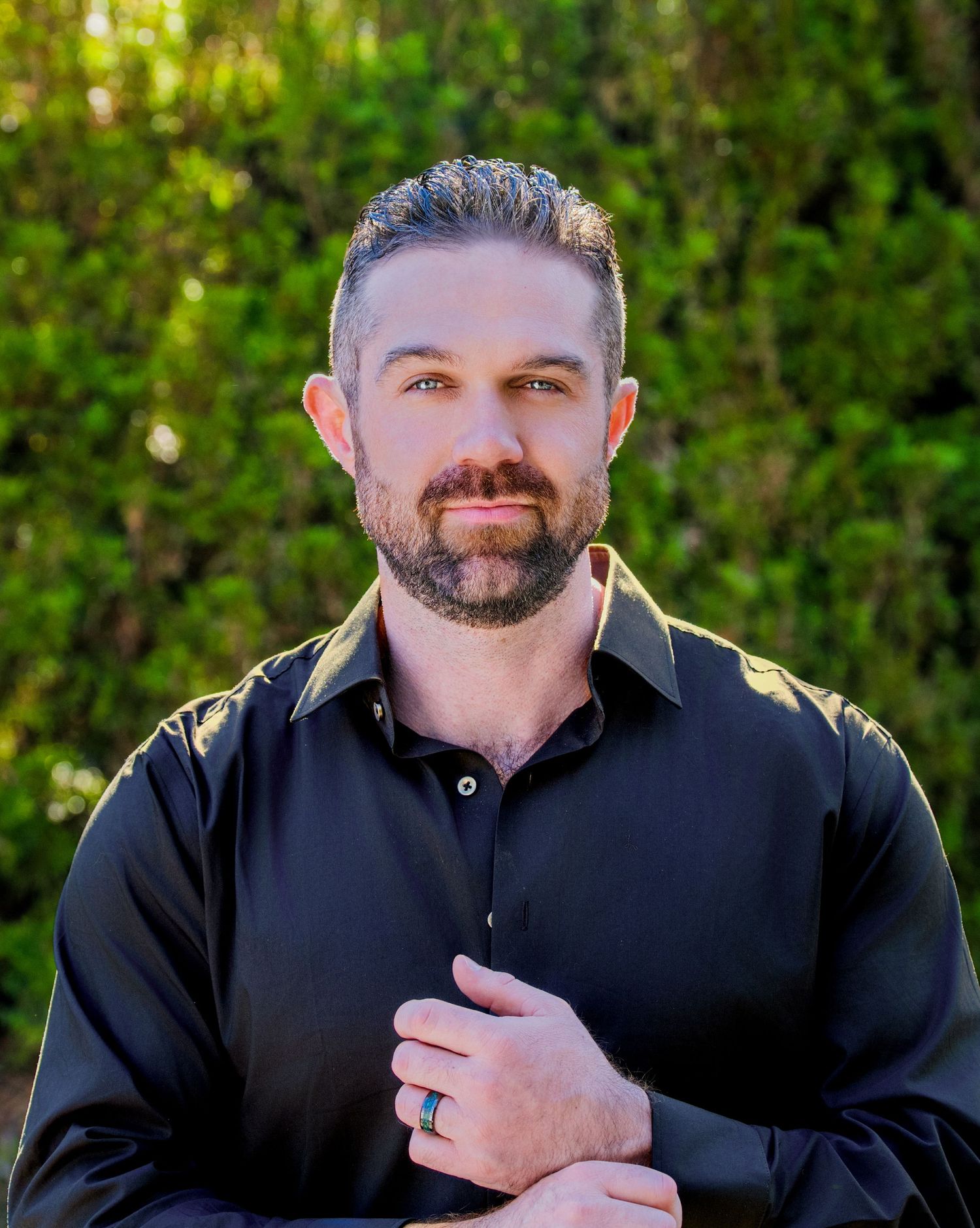 Professional man with beard in black shirt poses outdoors with green foliage background.
