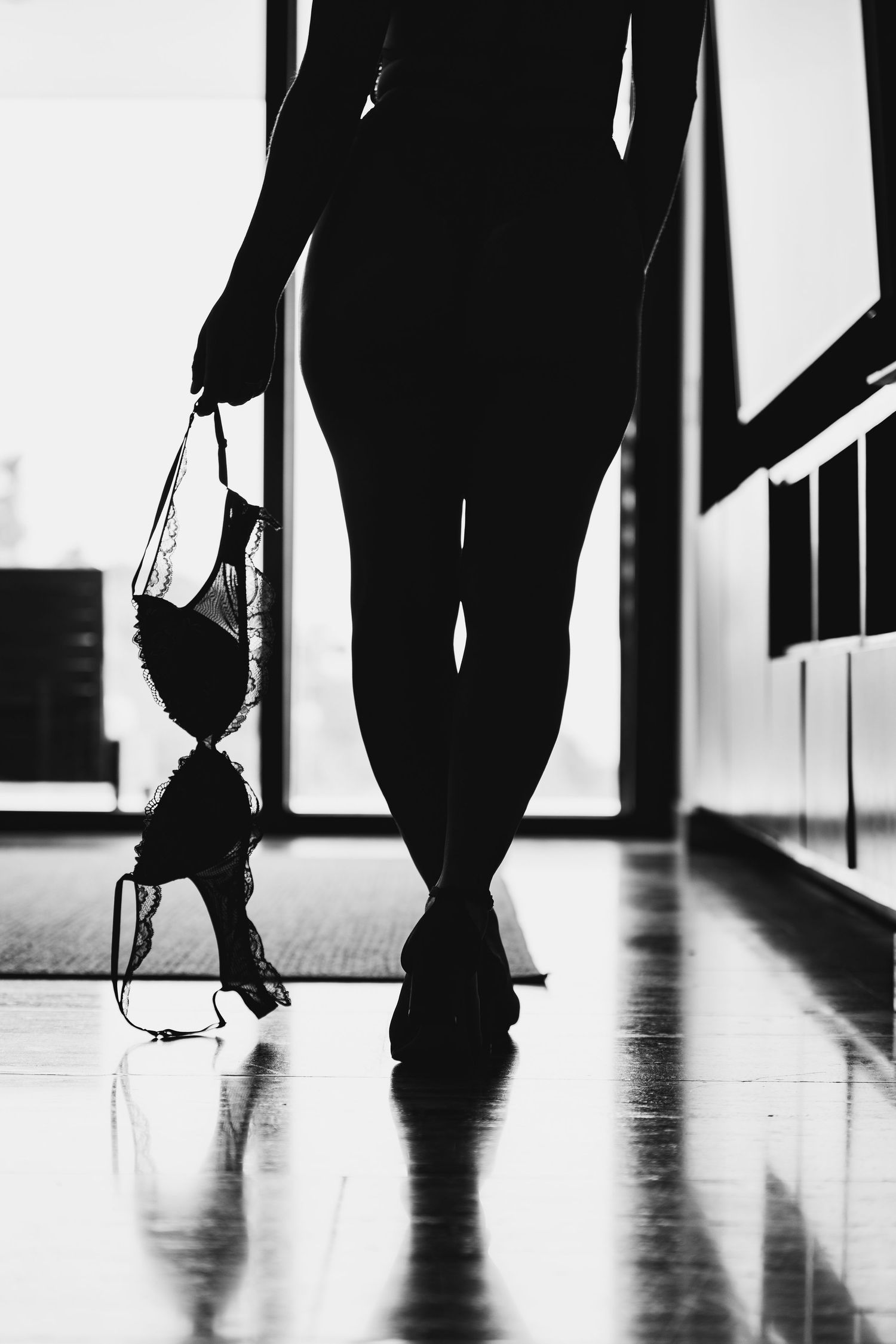 Black and white silhouette of woman walking down hallway holding lace bra in hand.