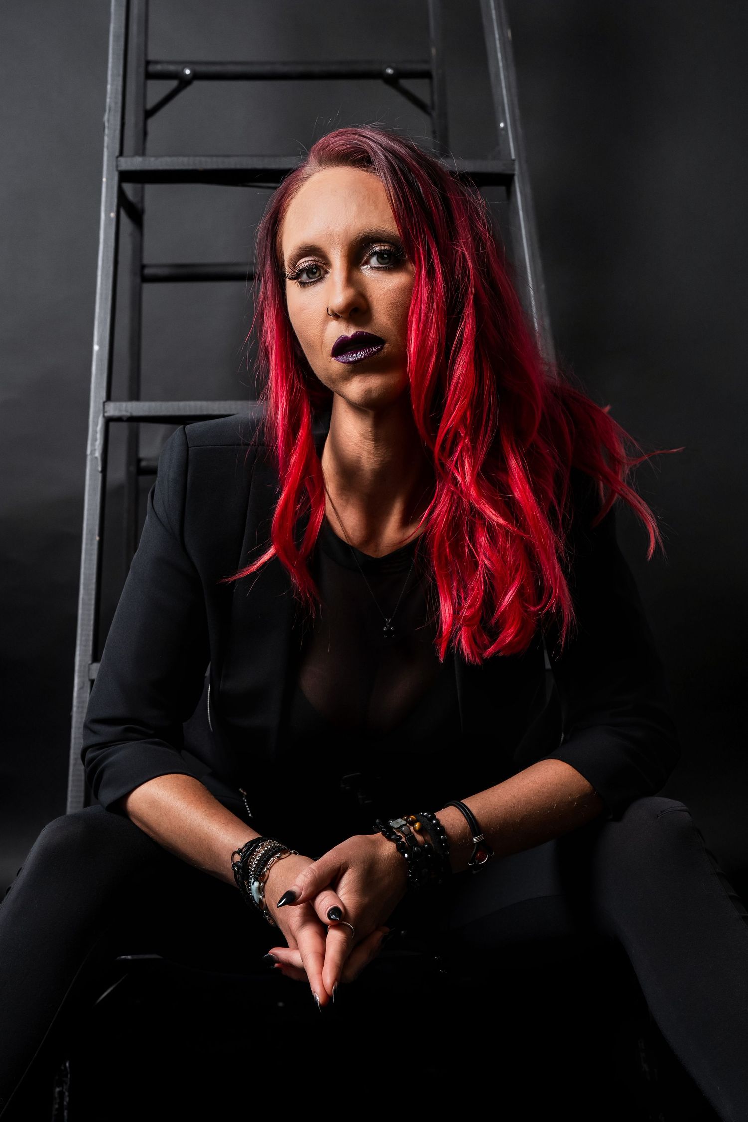 A woman with red hair and dark gothic makeup sits against a ladder in an all-black studio portrait.