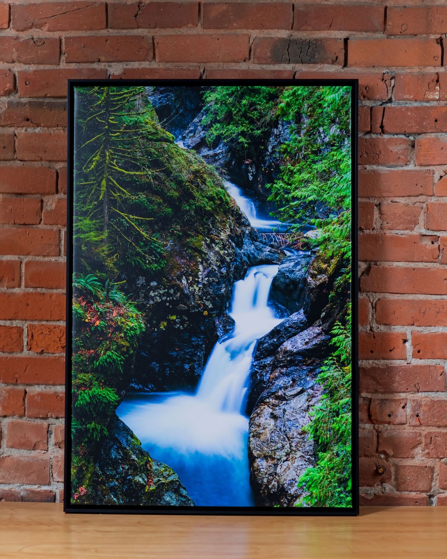 Framed waterfall photography print leaning against a brick wall, showcasing lush green moss-covered rocks and cascading blue water.