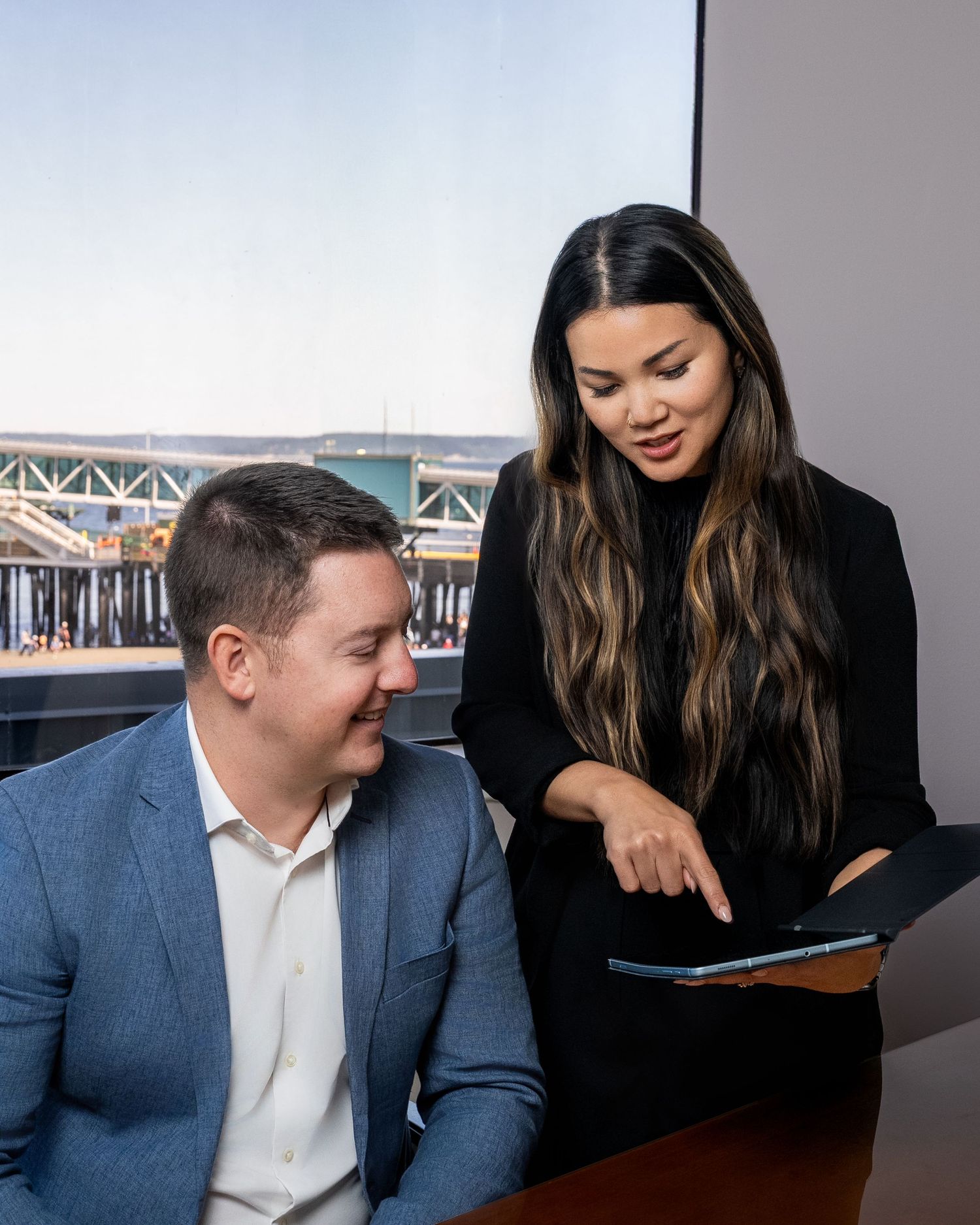 Man in blue suit and woman in black reviewing a tablet together in a modern office with waterfront view.