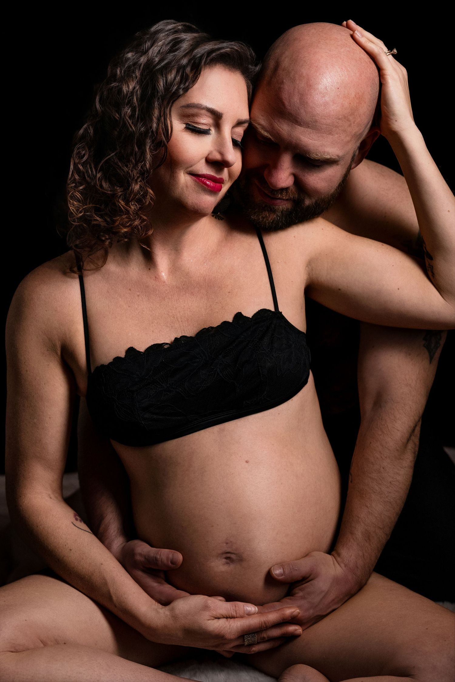 A pregnant couple shares an intimate moment, with the man tenderly cradling the woman's baby bump against a dark background.