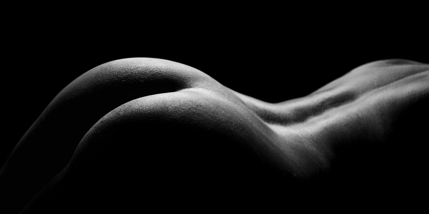 Black and white artistic nude bodyscape photograph highlighting curves of the human form against dark background.