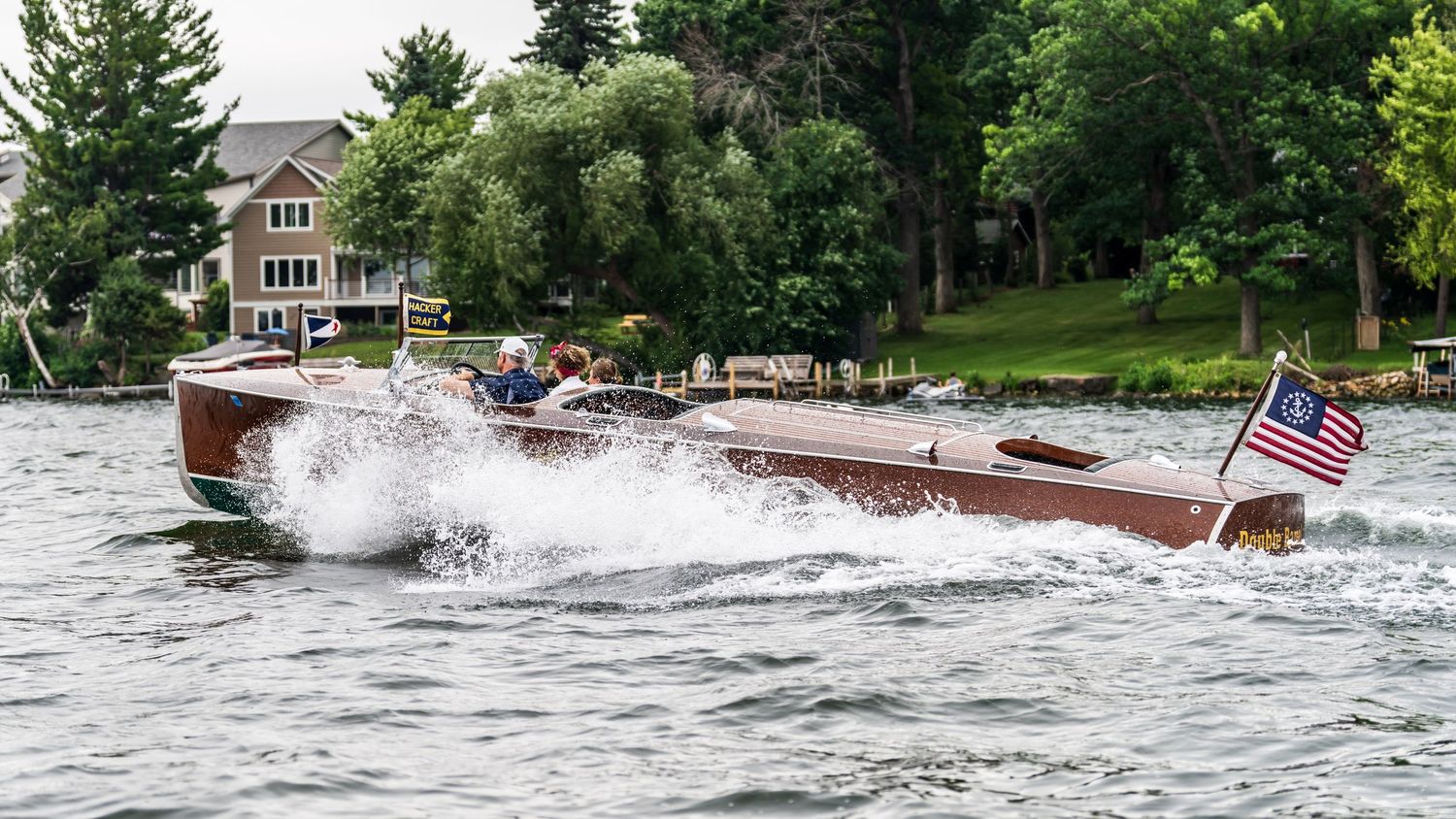 Classic wooden speedboat racing across a lake with an American flag, lush green trees and a house visible in the background.