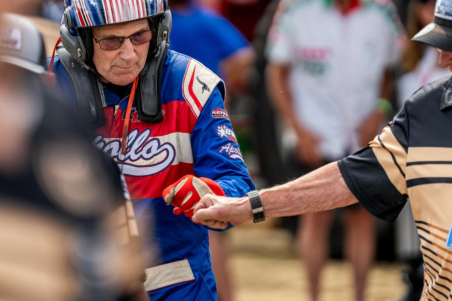 Elderly race car driver in patriotic suit shaking hands with another person at a motorsport event.