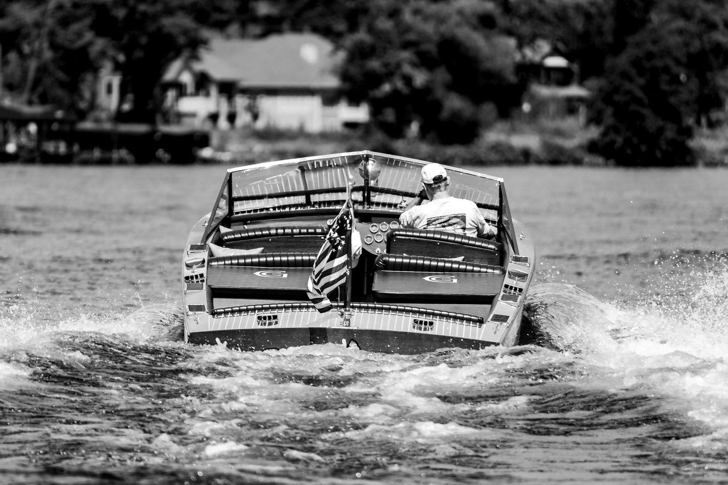 Black and white photo of vintage wooden speedboat racing across lake with American flag.