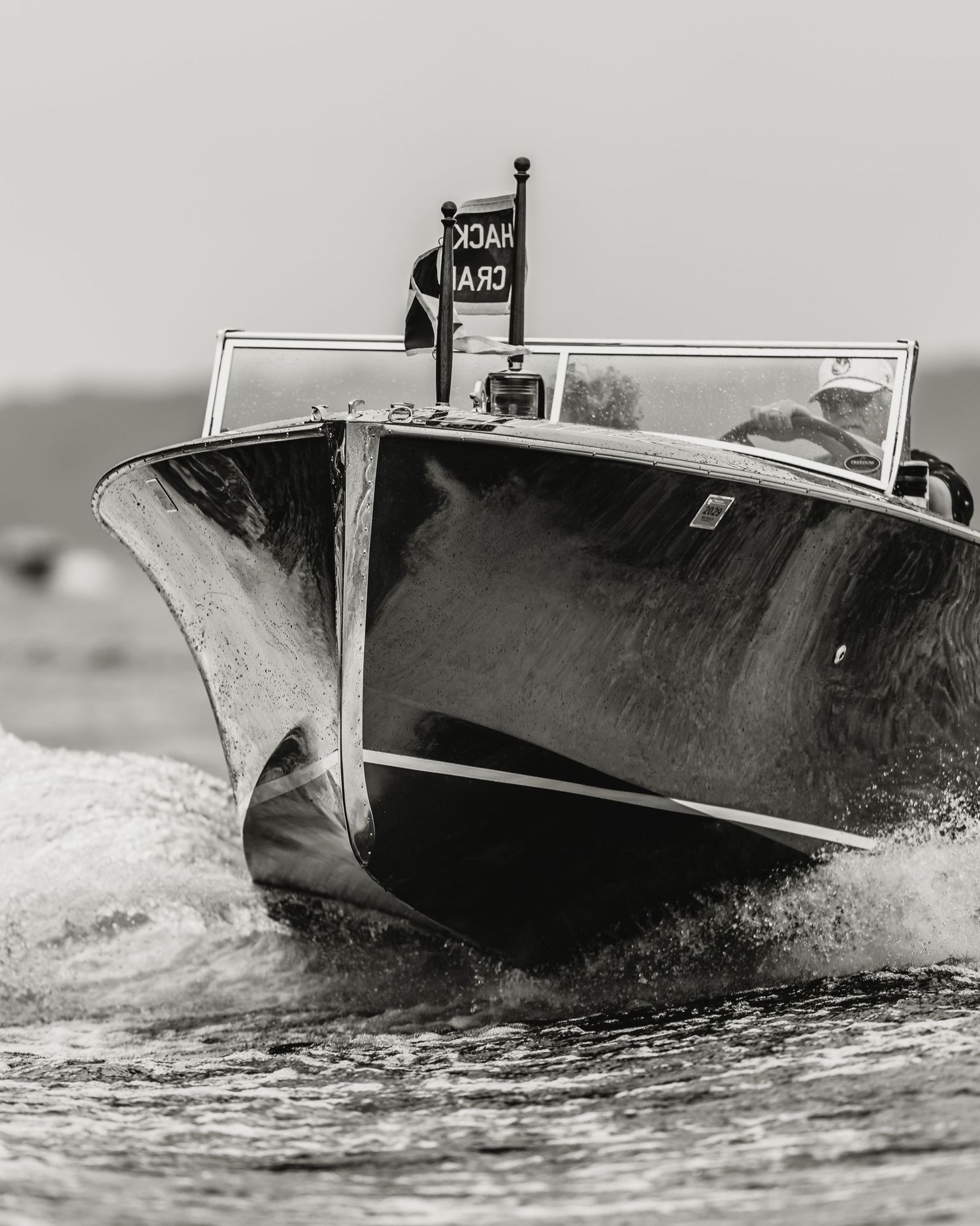 Black and white photo of a classic wooden speedboat racing across choppy water with a flag flying at the bow.