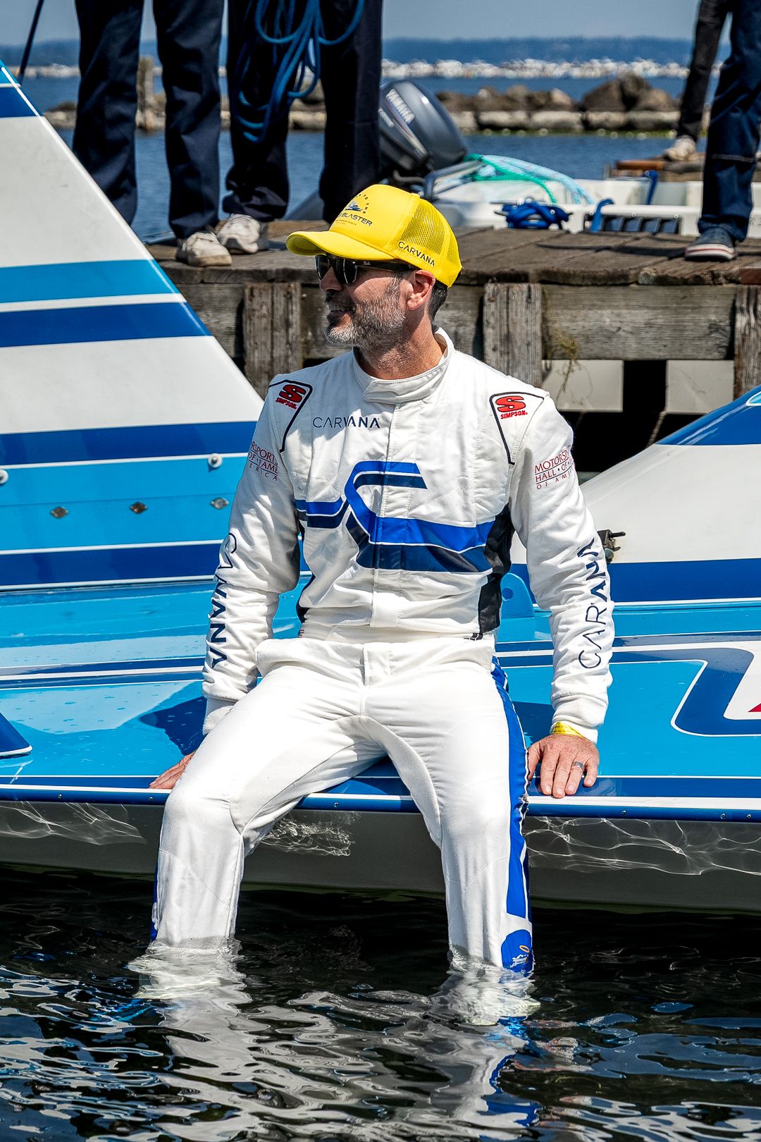 A male powerboat racer in white racing suit and yellow helmet sits on a boat hull with feet in water at a marina.